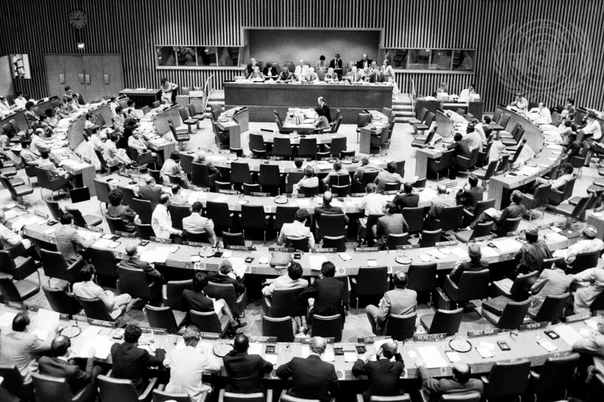 Overhead black and white photo of a United Nations assembly with delegates seated at curved desks.