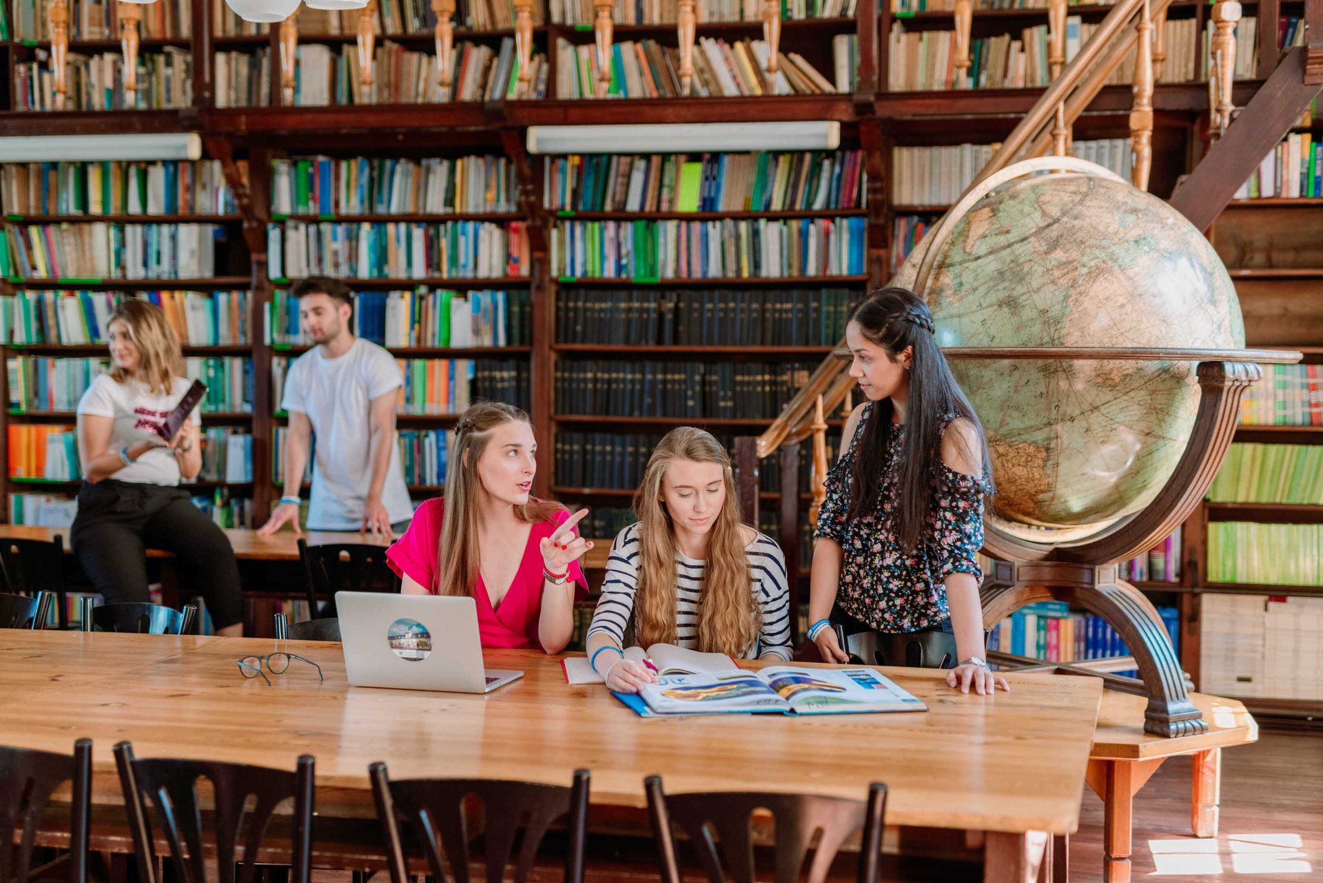 Young adults study and discuss at a large table in a vintage library with many books and a globe.