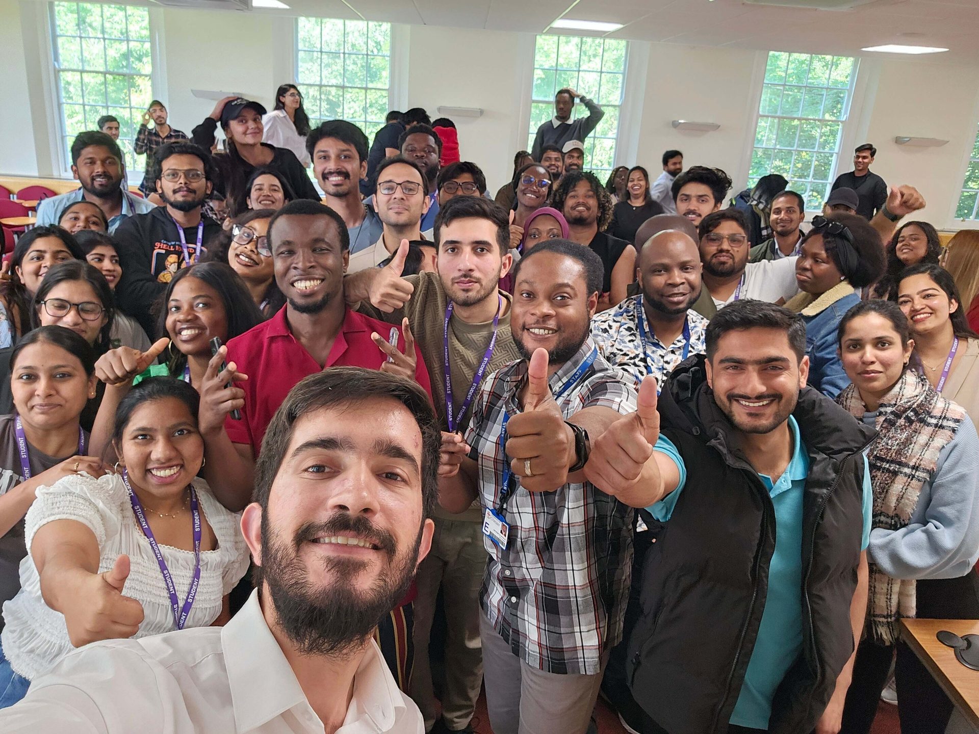 A large, diverse group of people smiling and giving thumbs up in a bright room with windows.