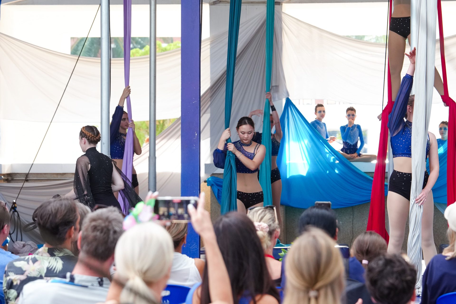 Aerialists perform on silks for an audience, some recording the event.