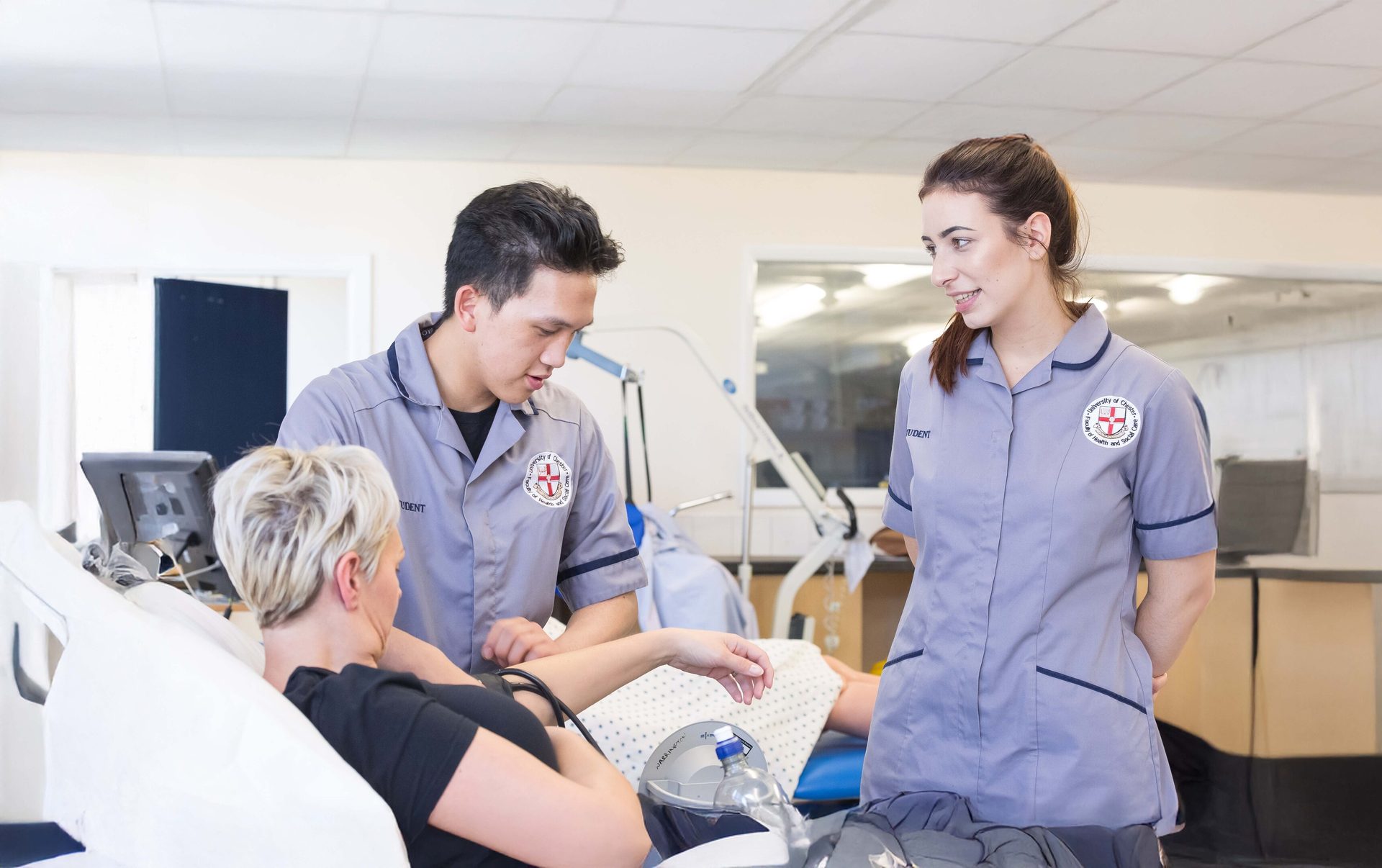 Two student nurses attend to a patient in a medical simulation lab, practicing patient care.