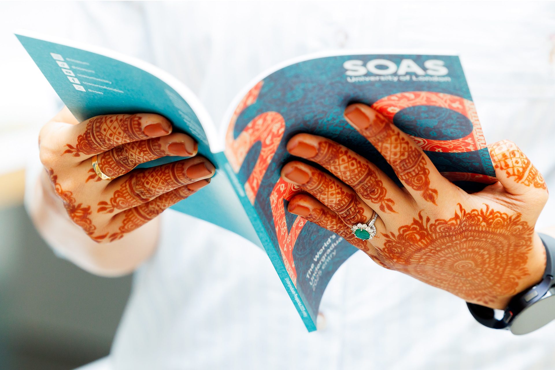 Hands with henna holding a SOAS University booklet, with rings and a watch.