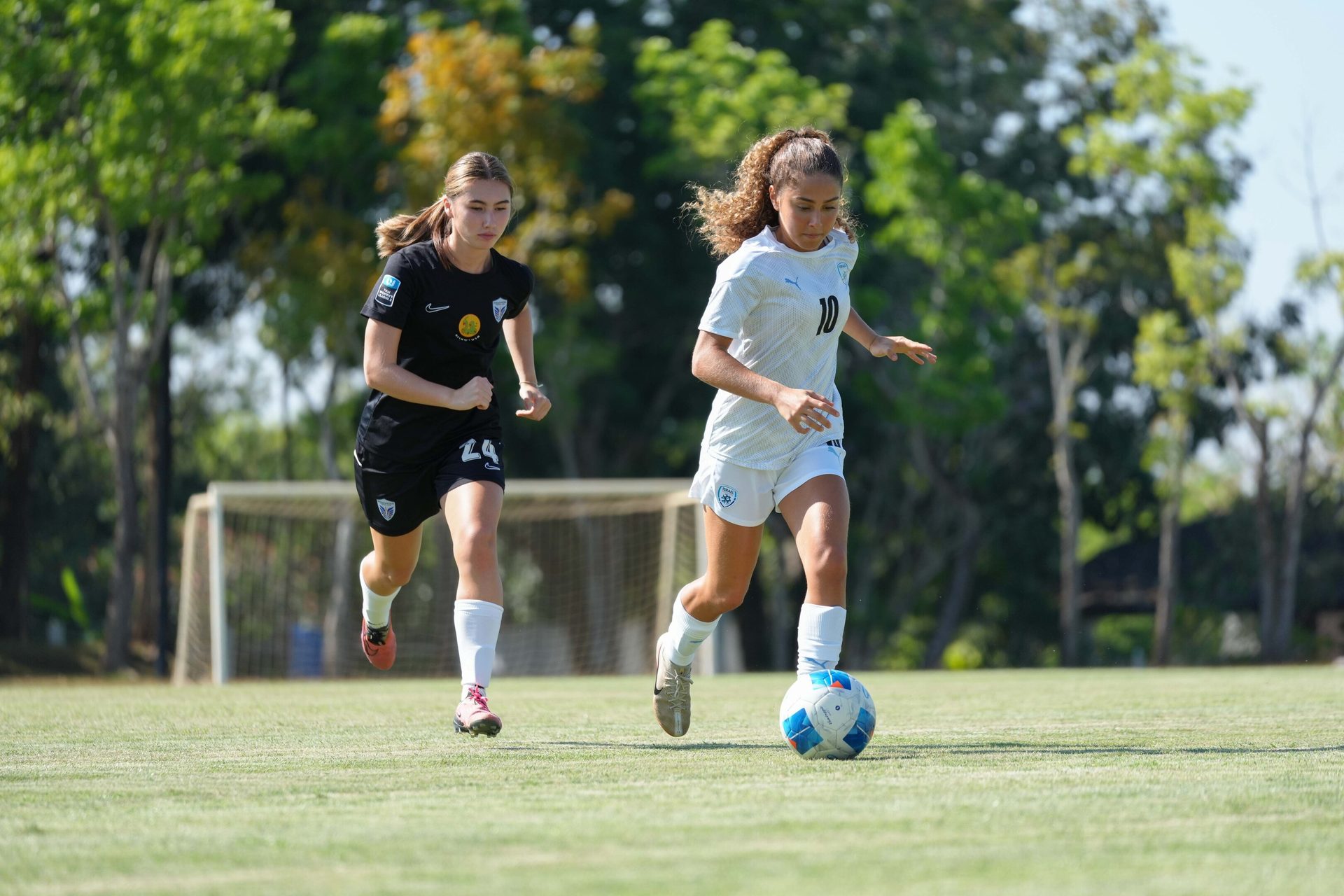 Two young female soccer players, one dribbling a ball, sprint across a green field.