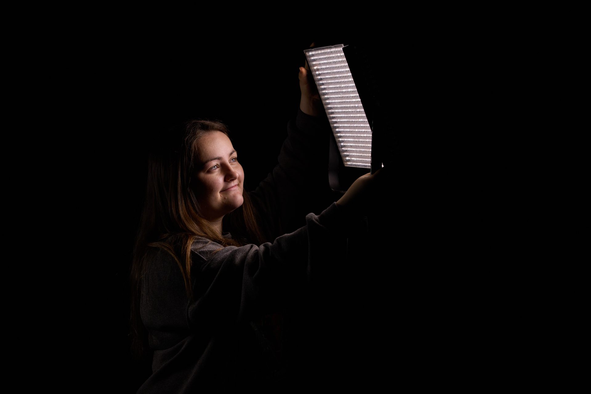A smiling woman holds an LED light panel that illuminates her face against a dark background.