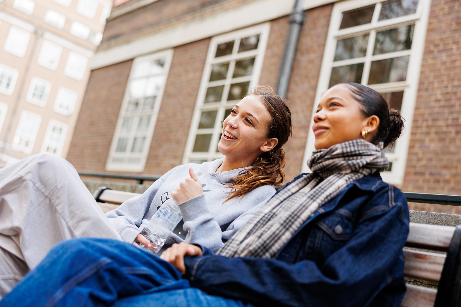 Two happy young women sit on a bench outdoors.