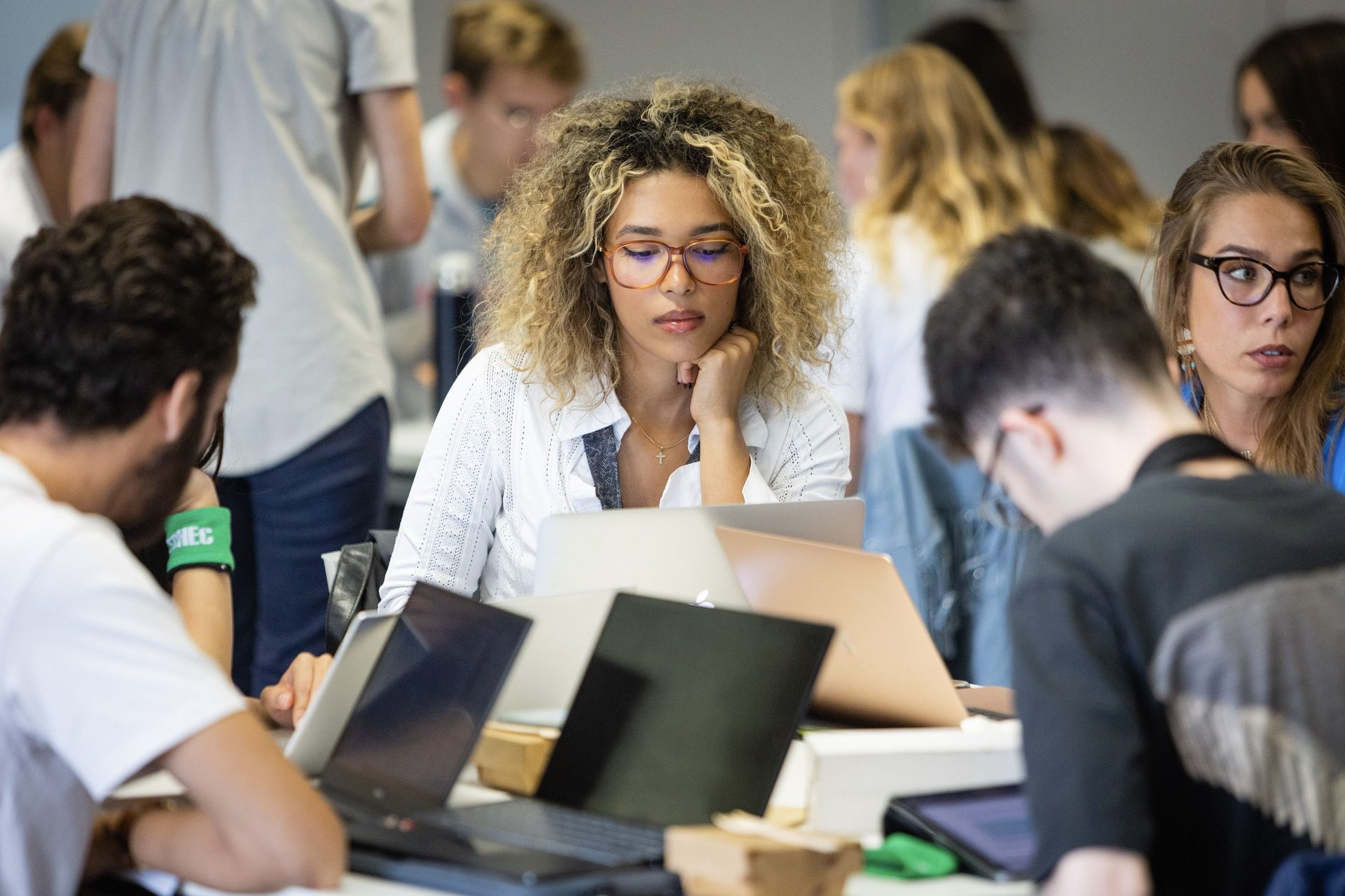 Woman with curly hair and glasses focused on laptop among students working in a classroom.