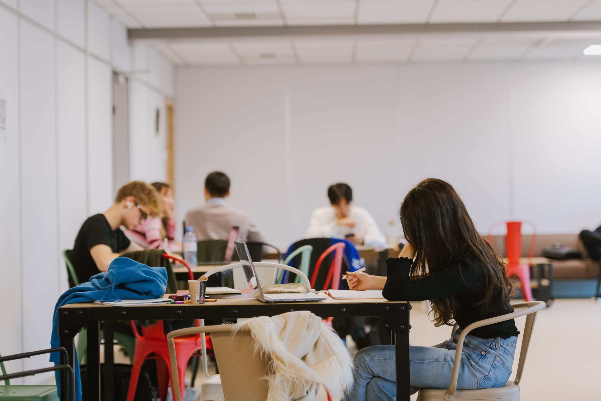 Students studying at tables in a bright room, a girl writing in the foreground.