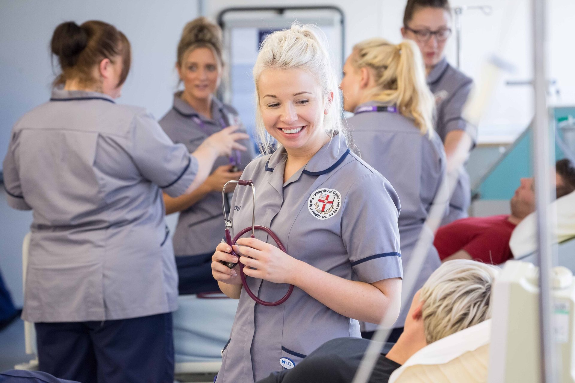 A smiling nurse in grey scrubs holds a stethoscope, with other nurses and patients in a hospital.