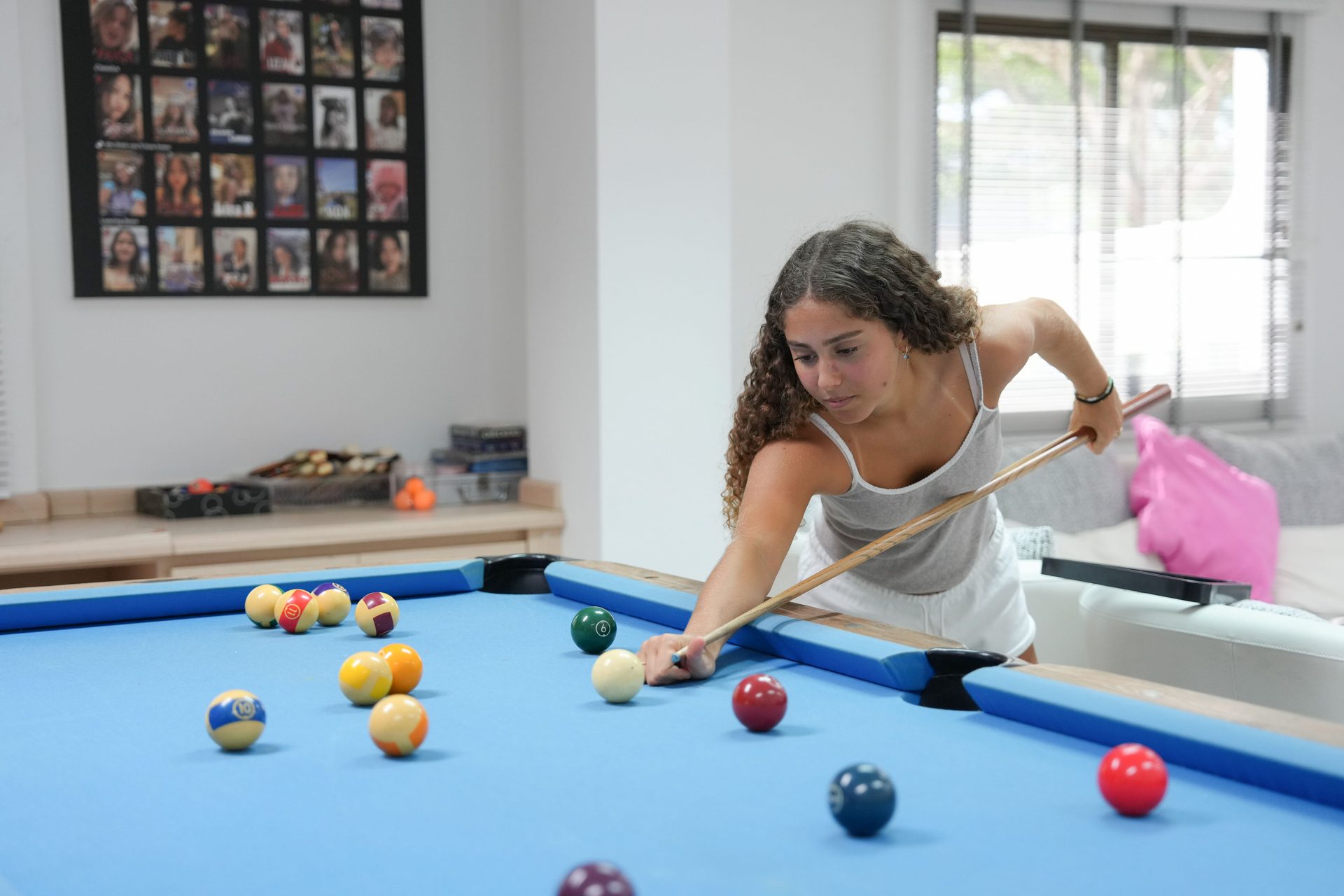 A girl with curly hair plays billiards on a bright blue pool table, aiming her shot.