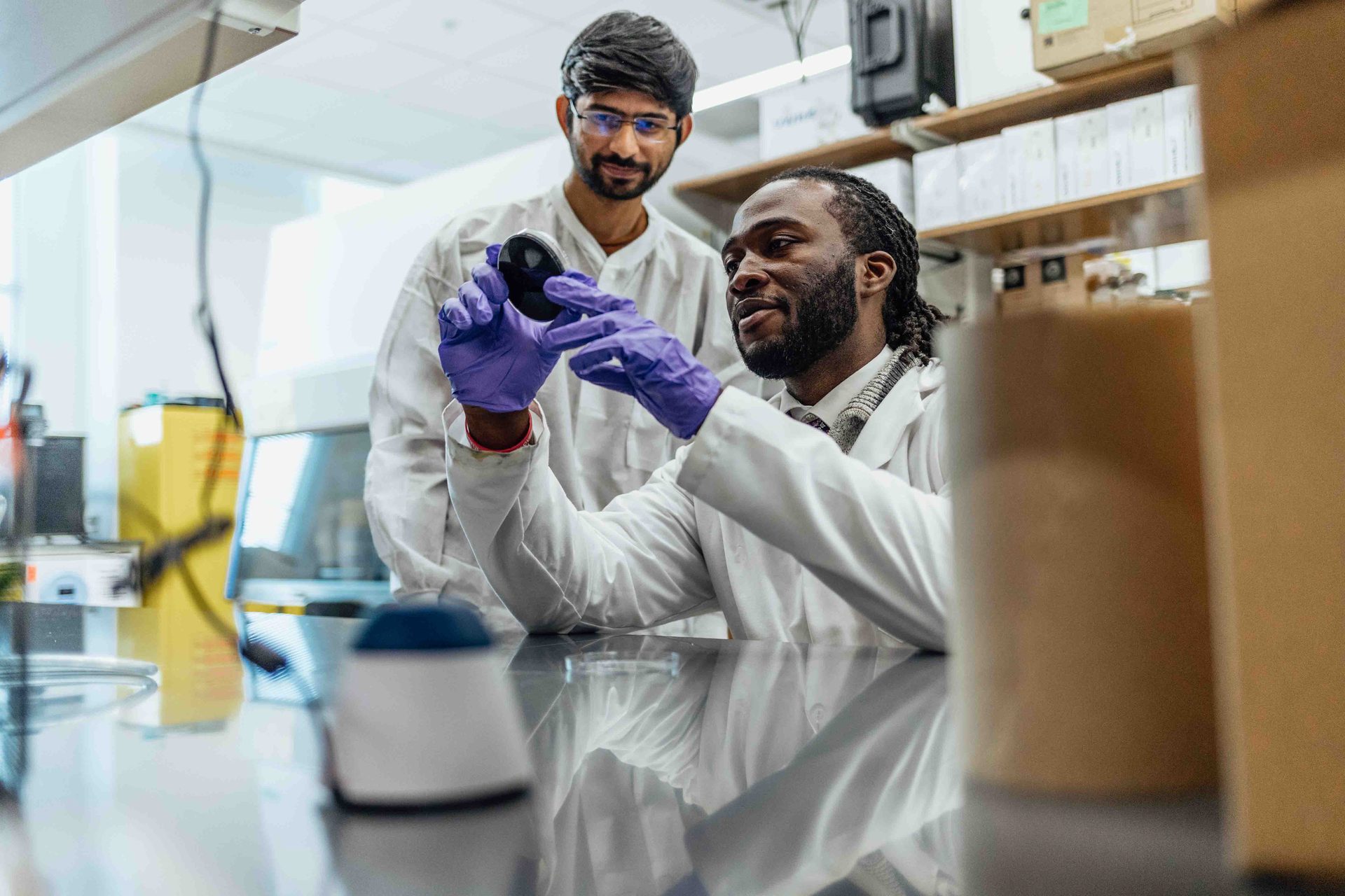 Two scientists in lab coats and gloves examine a sample in a laboratory setting.