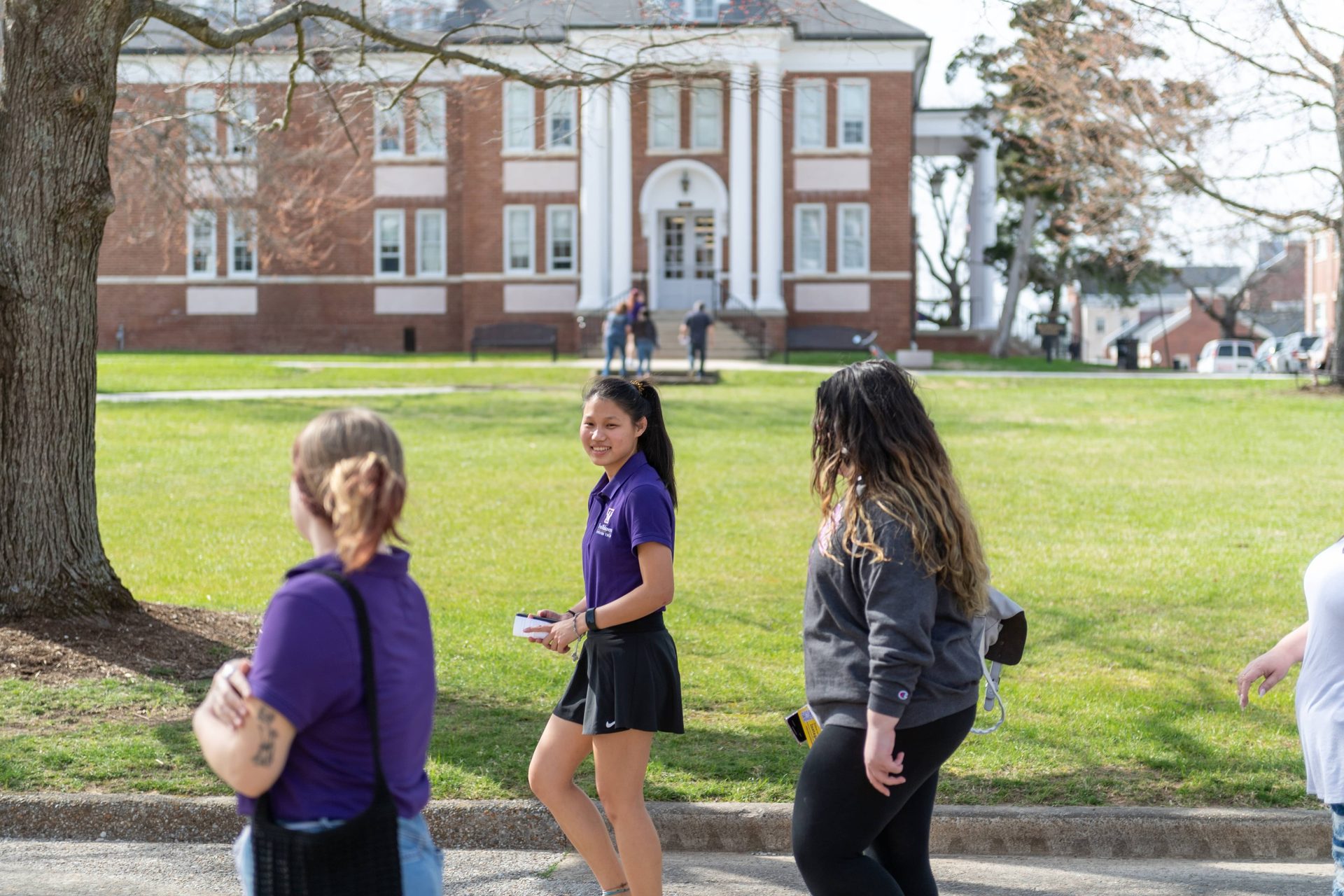 Smiling student in purple walks on campus with others, brick building in background.