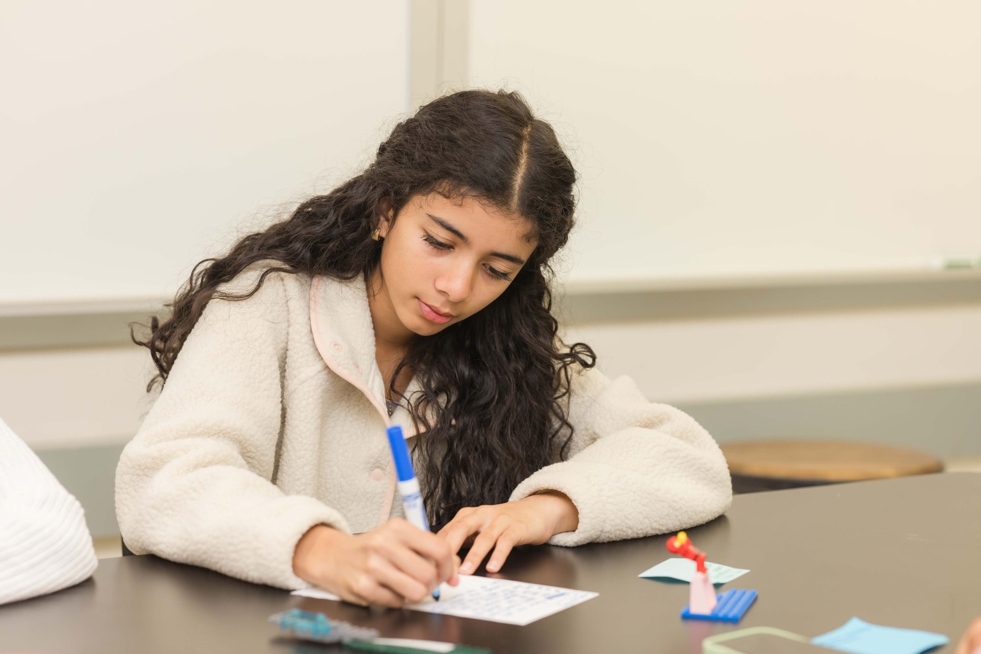 A young girl with long, curly dark hair intently writes on paper with a blue marker at a table.