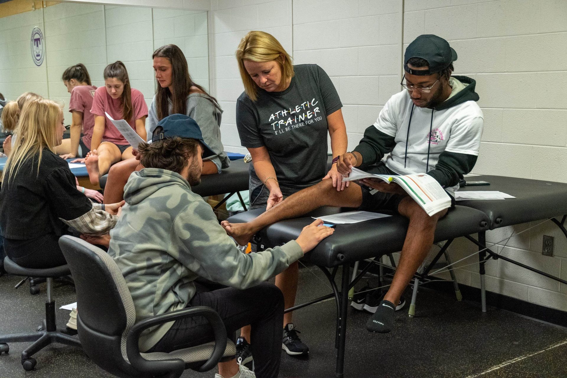 Athletic trainer examines a man's leg in a classroom with other students present.