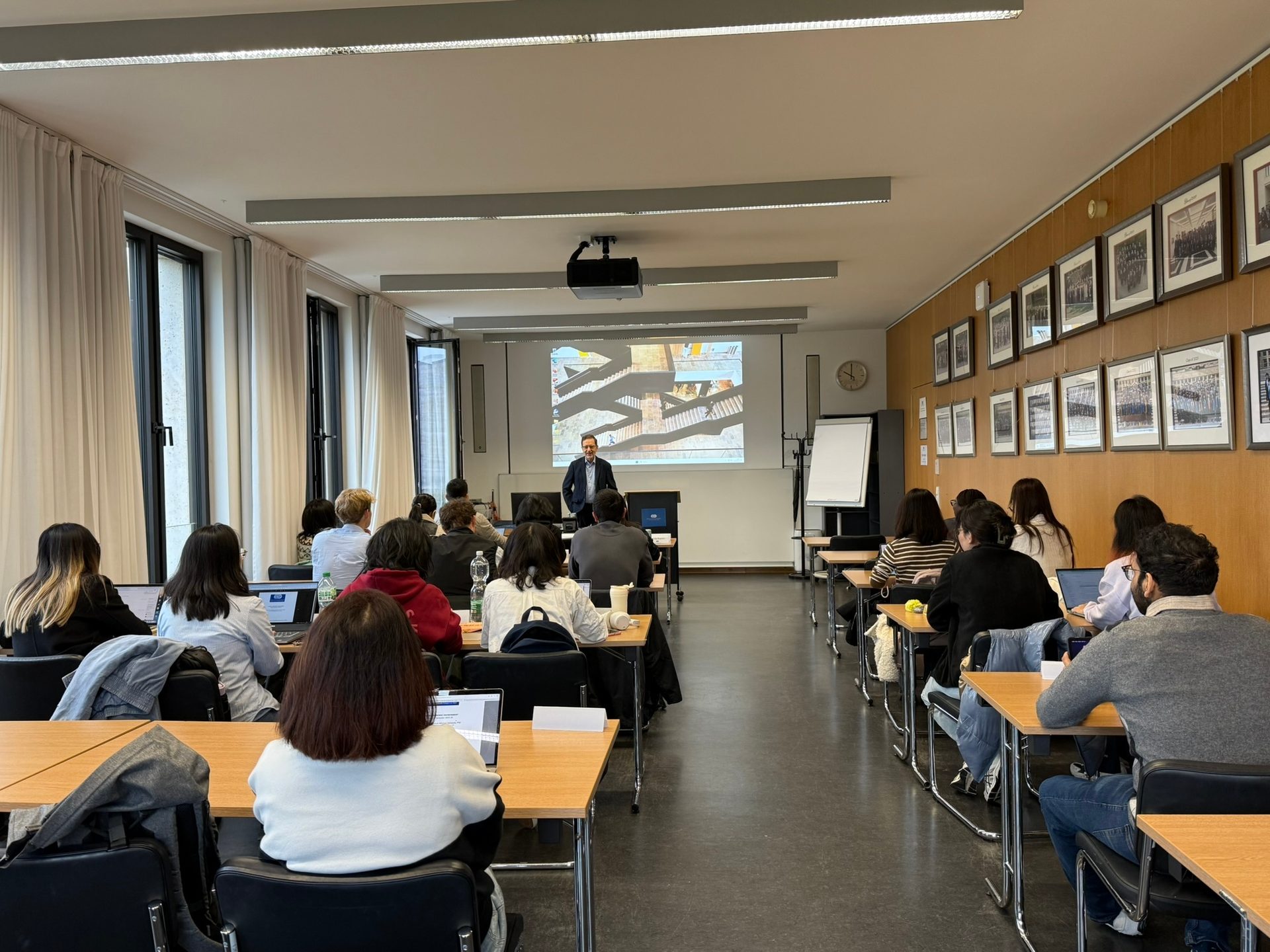 Lecturer presents to students in a classroom with a projector, windows, and framed pictures.