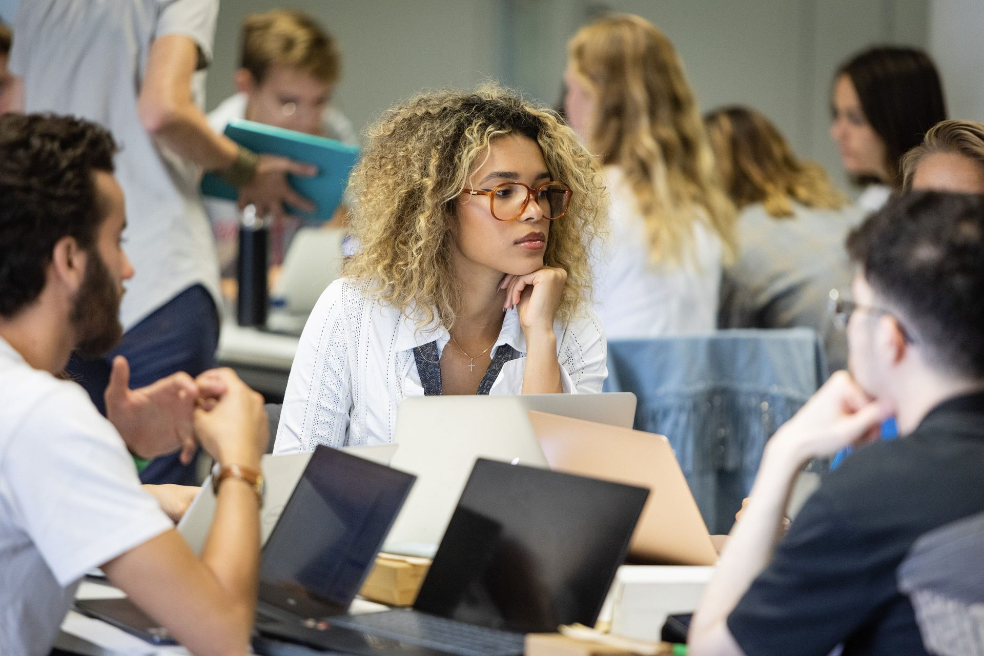 Pensive young woman with curly hair and glasses, hand on chin, among laptops and peers.