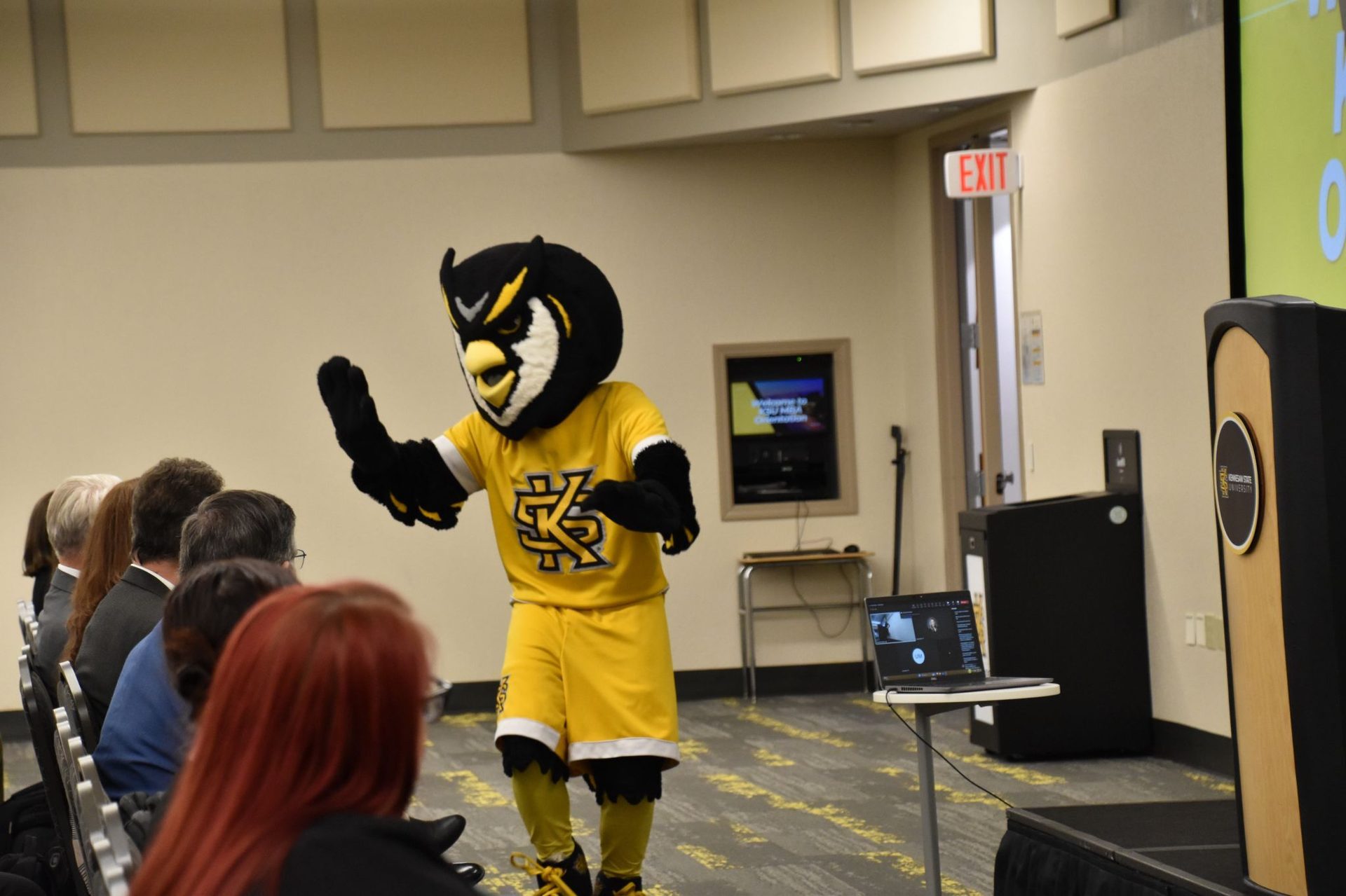 Owl mascot (Kennesaw State University) standing in an event hall with an audience and podium.