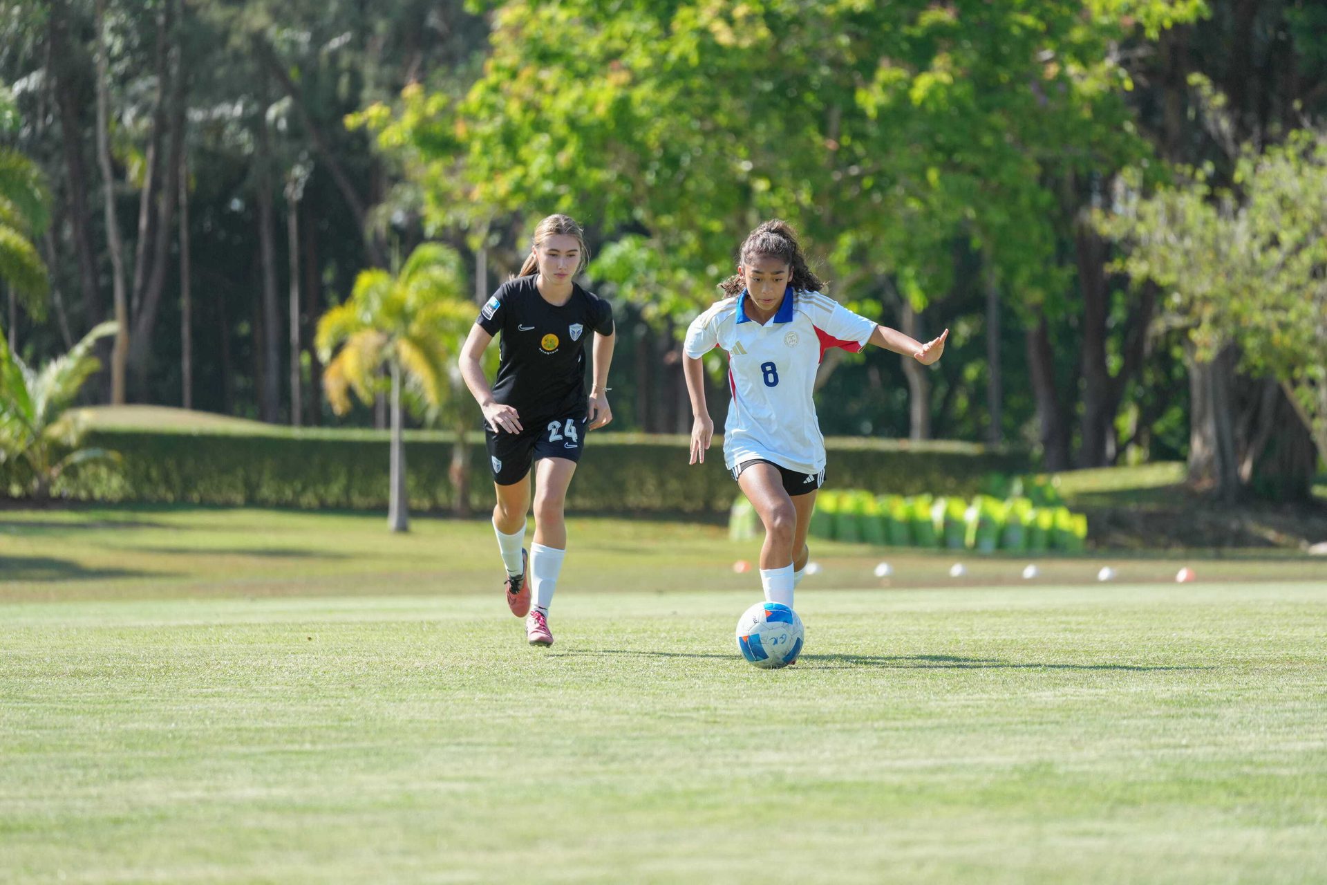 Two young women playing soccer on a grassy field, one dribbling the ball, the other running behind.