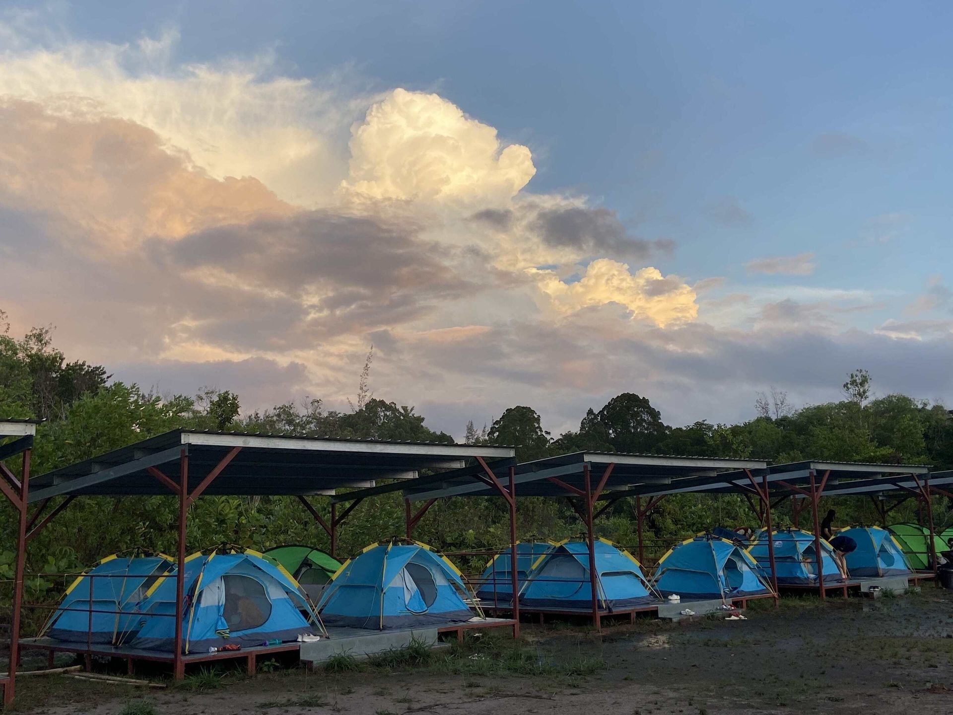 Blue tents under metal roofs at a campsite with trees and a dramatic cloudy sky.