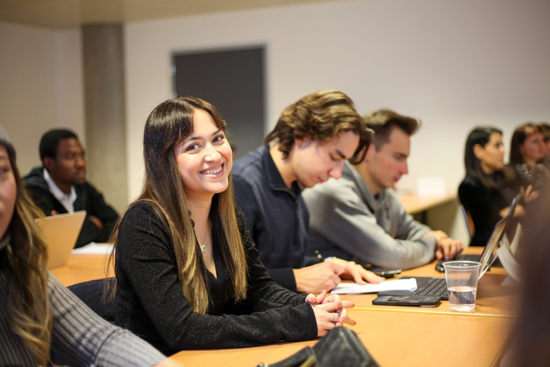 A smiling young woman looks at the camera from a classroom table, surrounded by other students.