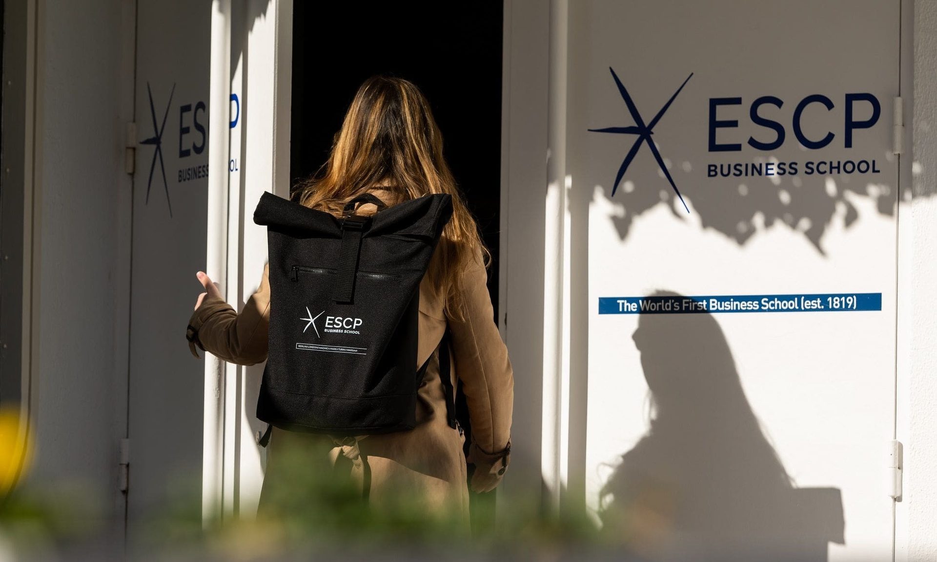 Student with ESCP Business School backpack entering door with 'The World's First Business School (est. 1819)'.