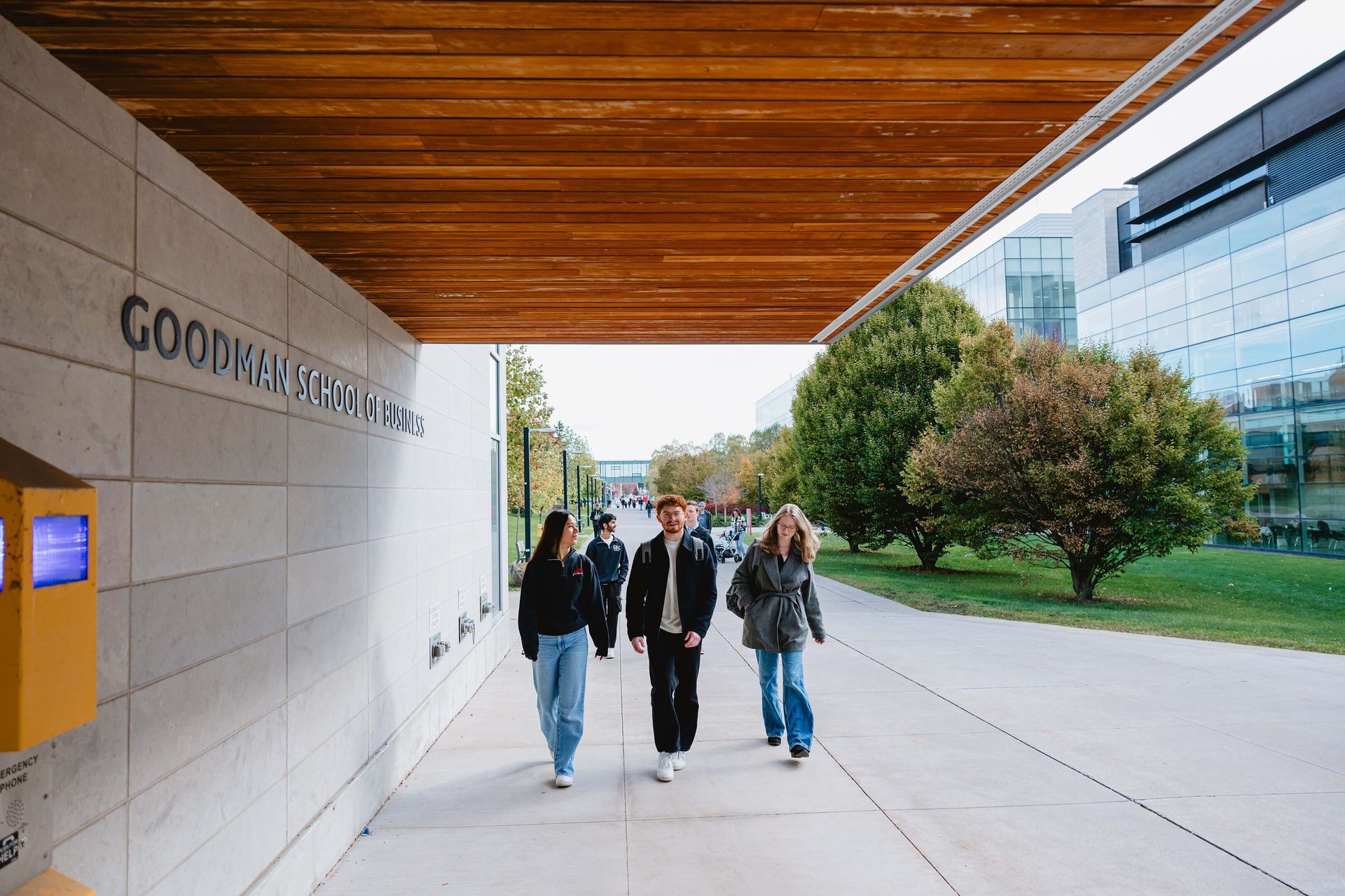 Three people walk past "Goodman School of Business" sign on a campus path.