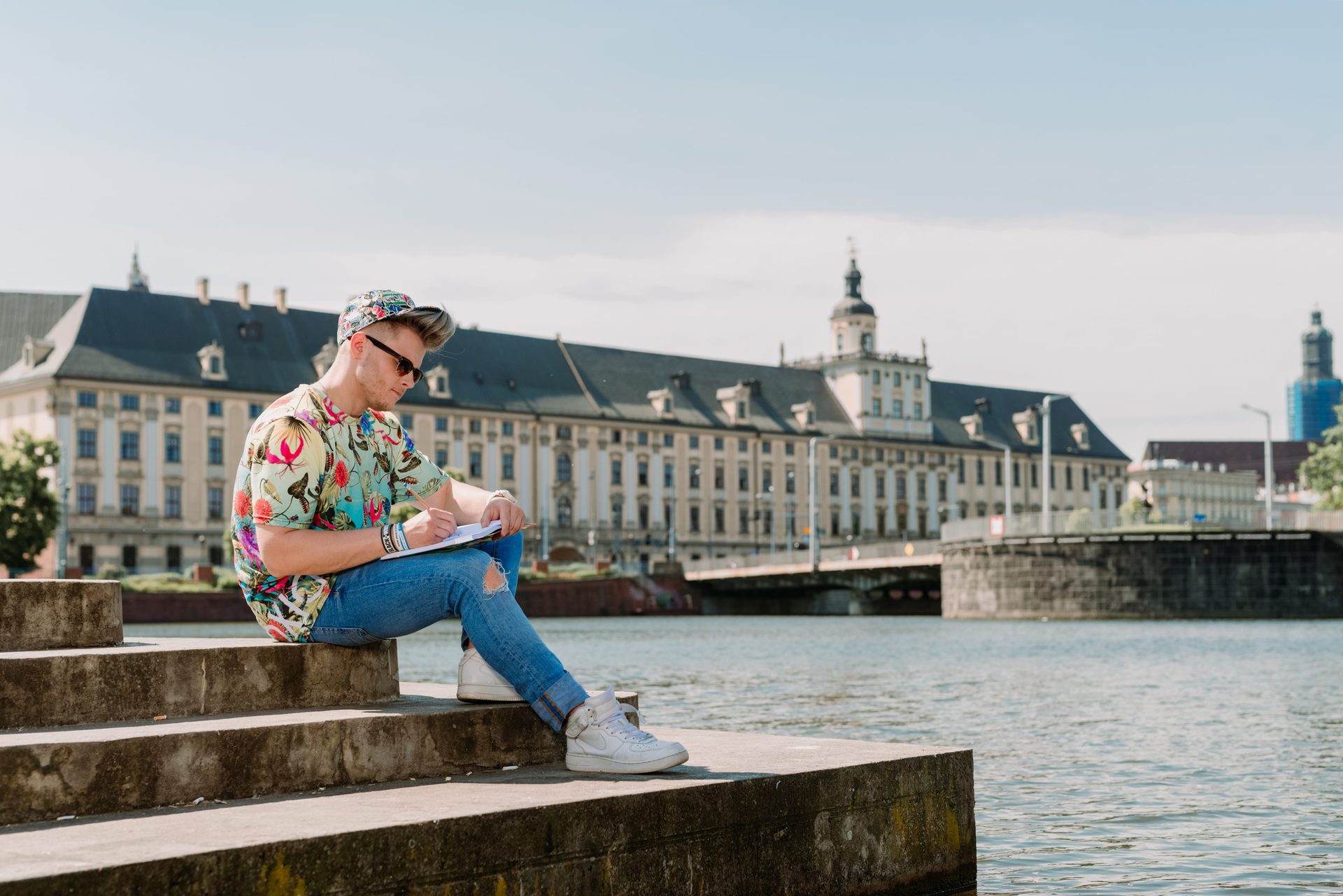 Young man in a vibrant shirt sits by a river, writing in a notebook, with a grand historic building behind him.