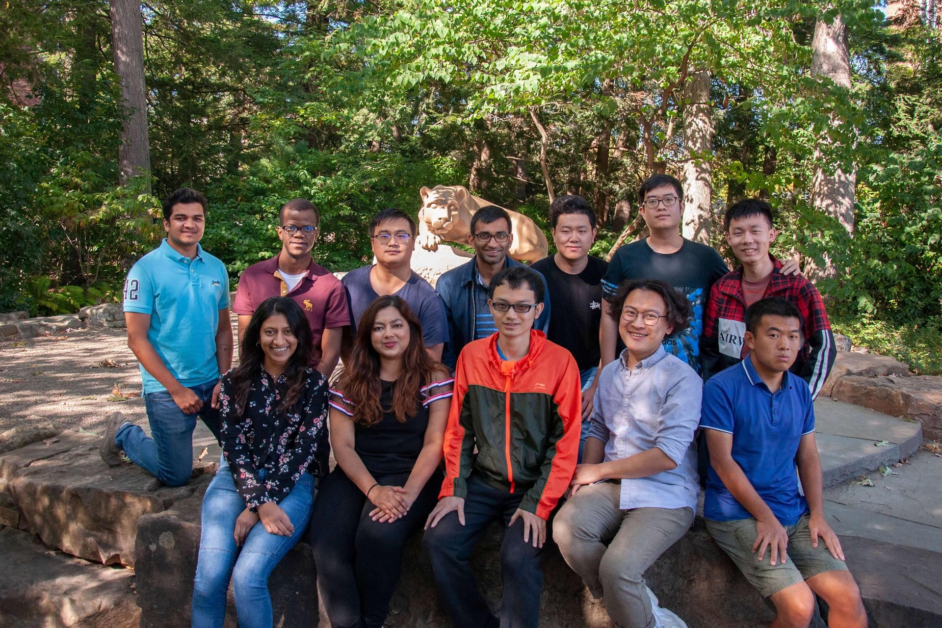 A diverse group of 14 smiling young adults poses outdoors with a cougar statue behind them.