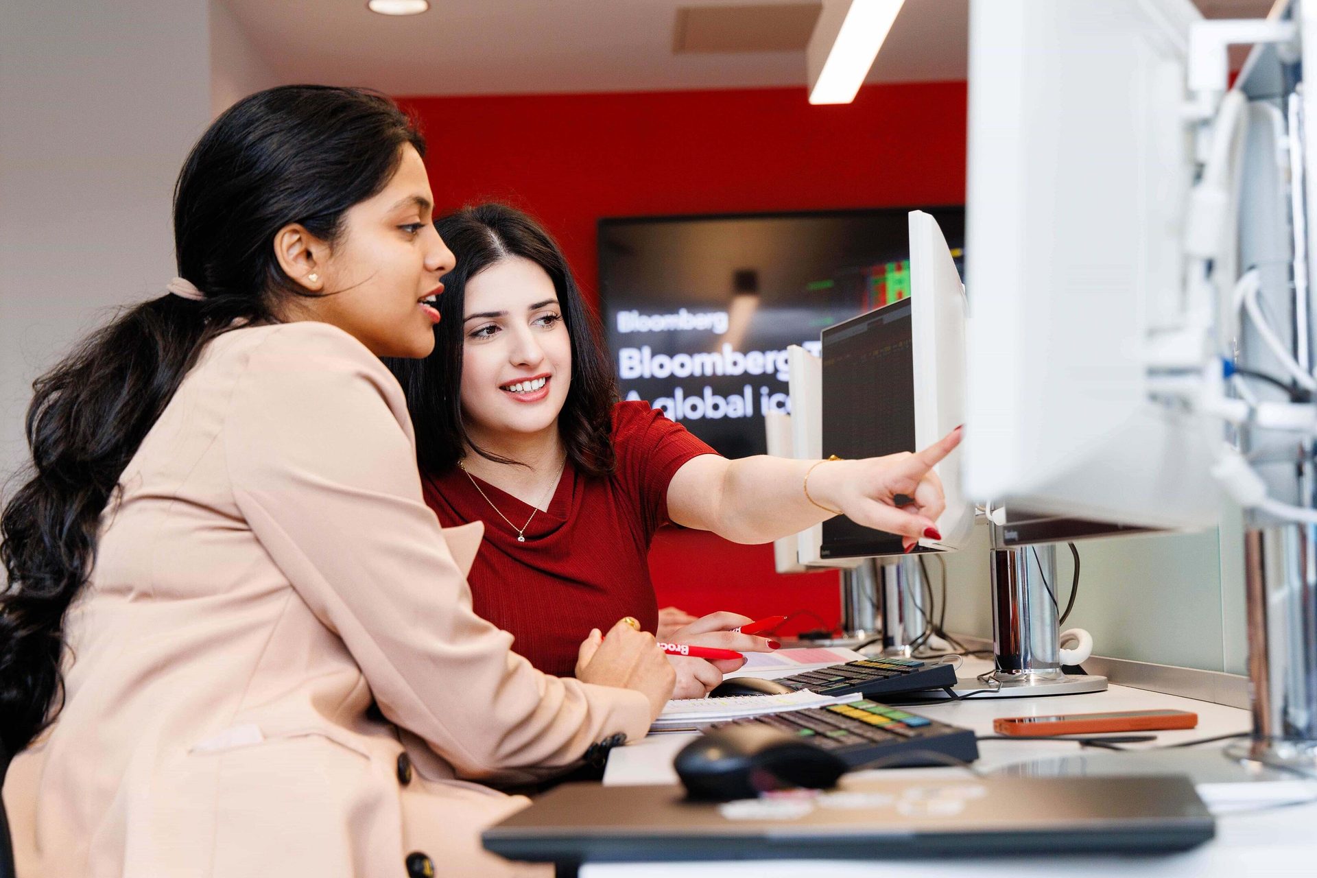 Two women collaborate at a Bloomberg terminal, one pointing at the screen.