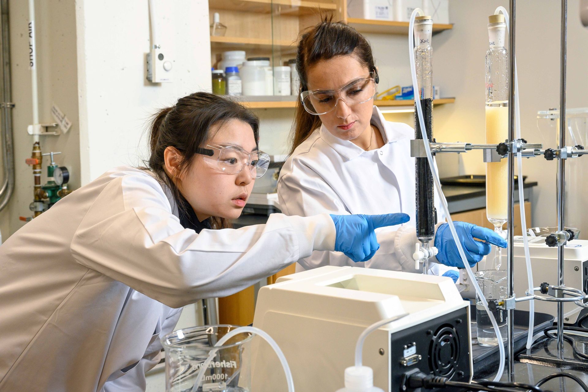 Two scientists in safety gear pointing at lab equipment during an experiment.