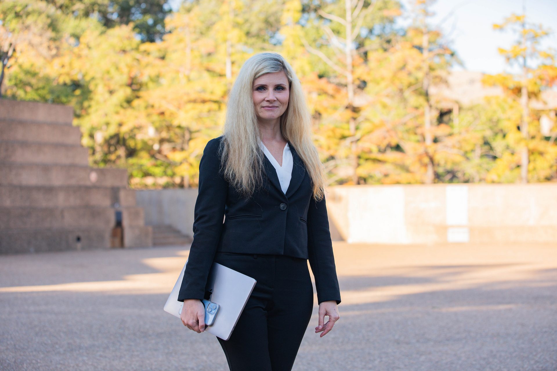 A blonde woman in a black business suit holds a laptop, standing outdoors with trees in the background.