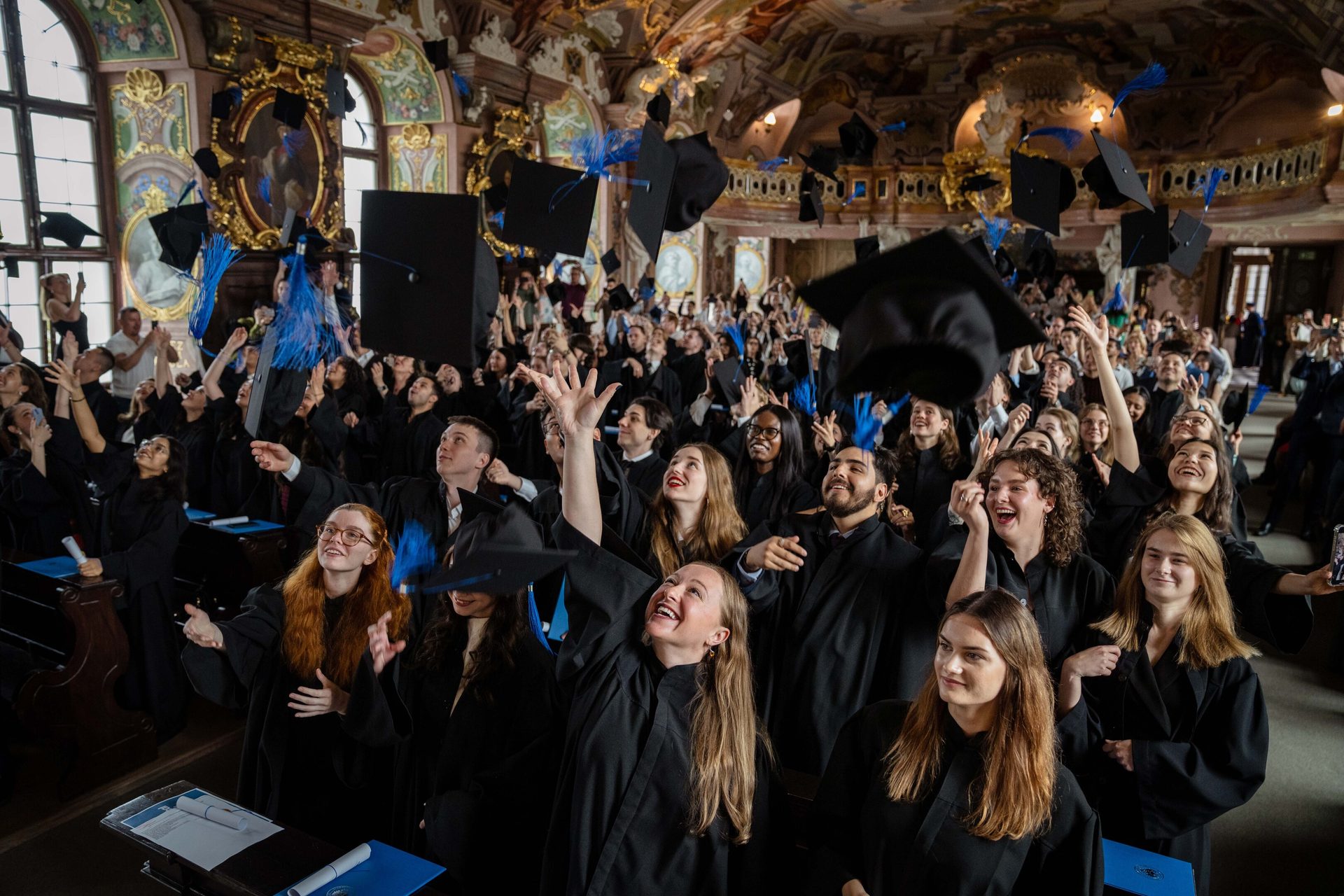 Graduates in academic robes joyfully throw their caps in a grand hall, celebrating their achievement.