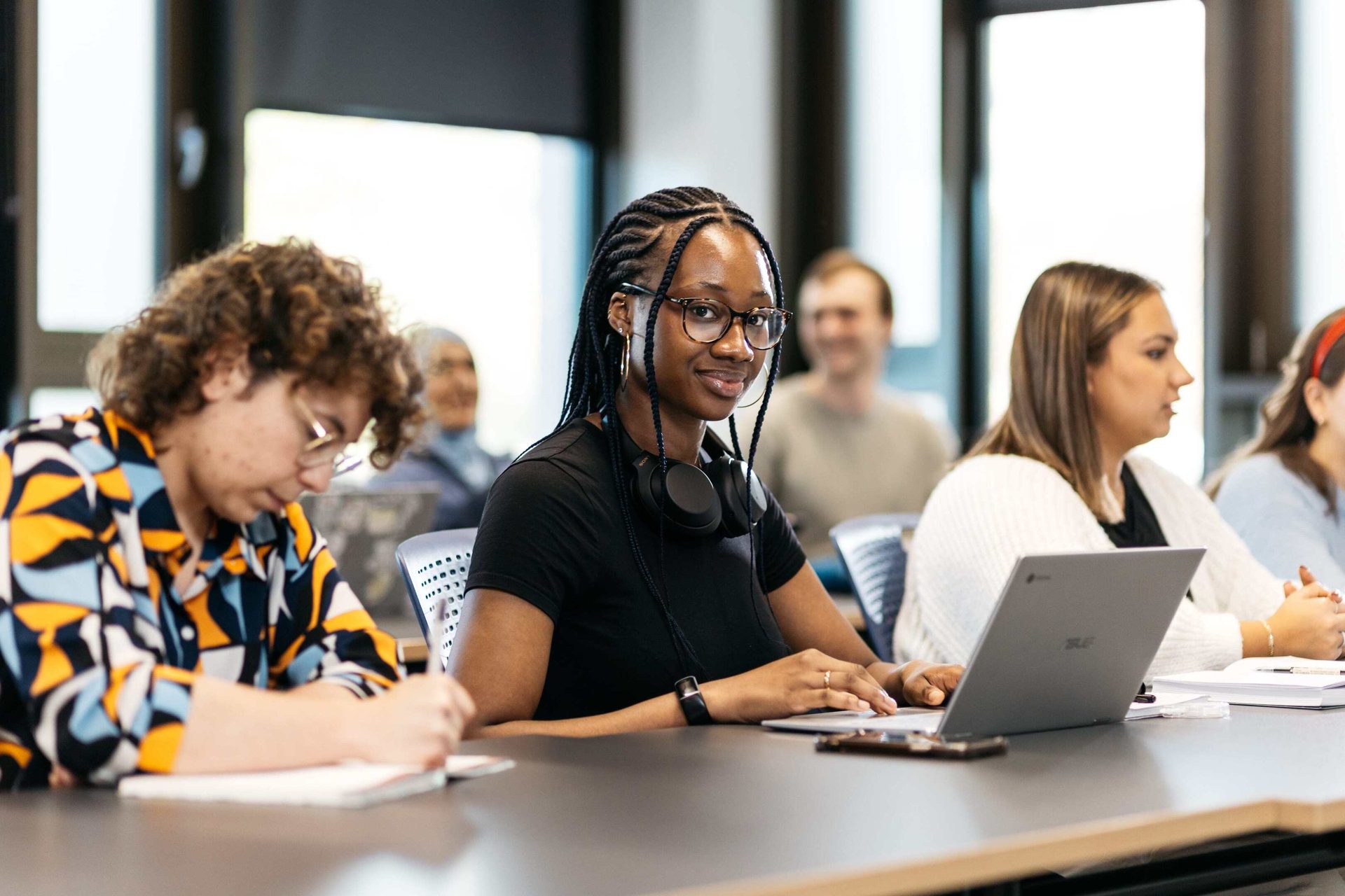 Diverse young adults in a classroom. A Black woman with braids and glasses smiles, typing on a laptop.