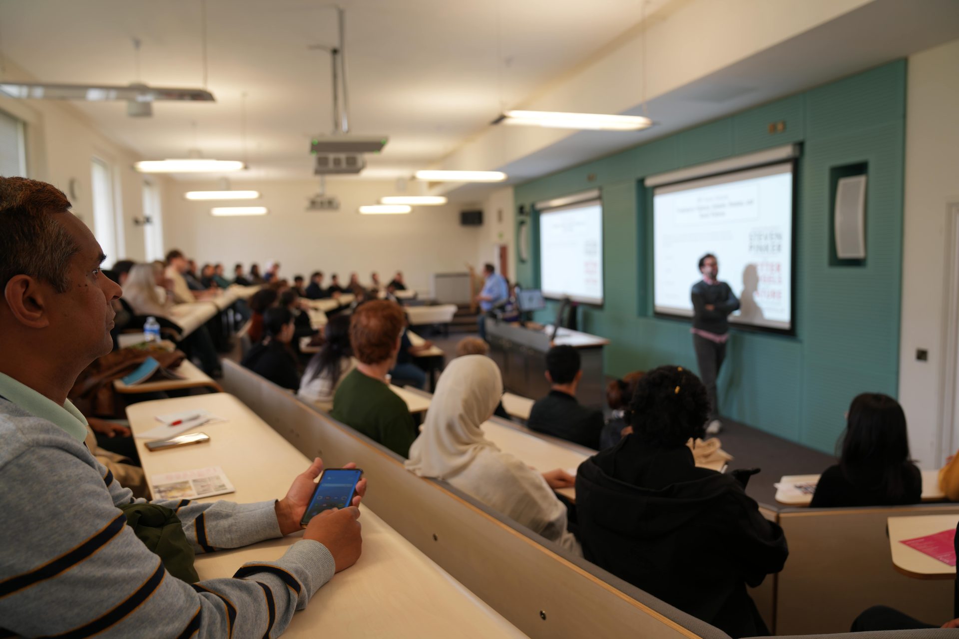 Classroom scene: student in foreground with phone, lecturer presents to class.