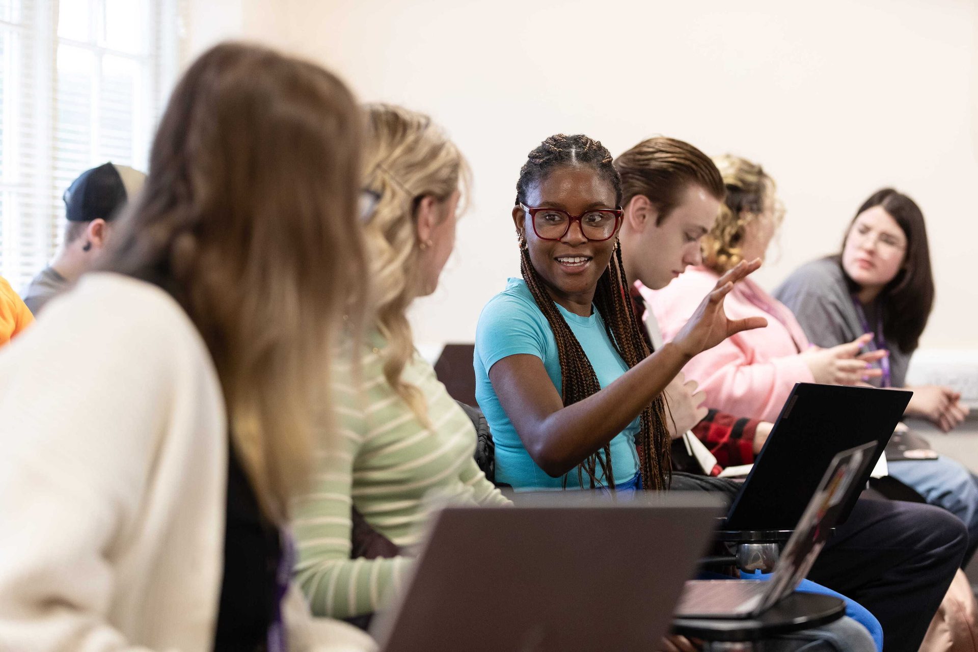 A smiling student gestures while speaking in a classroom surrounded by other students and laptops.