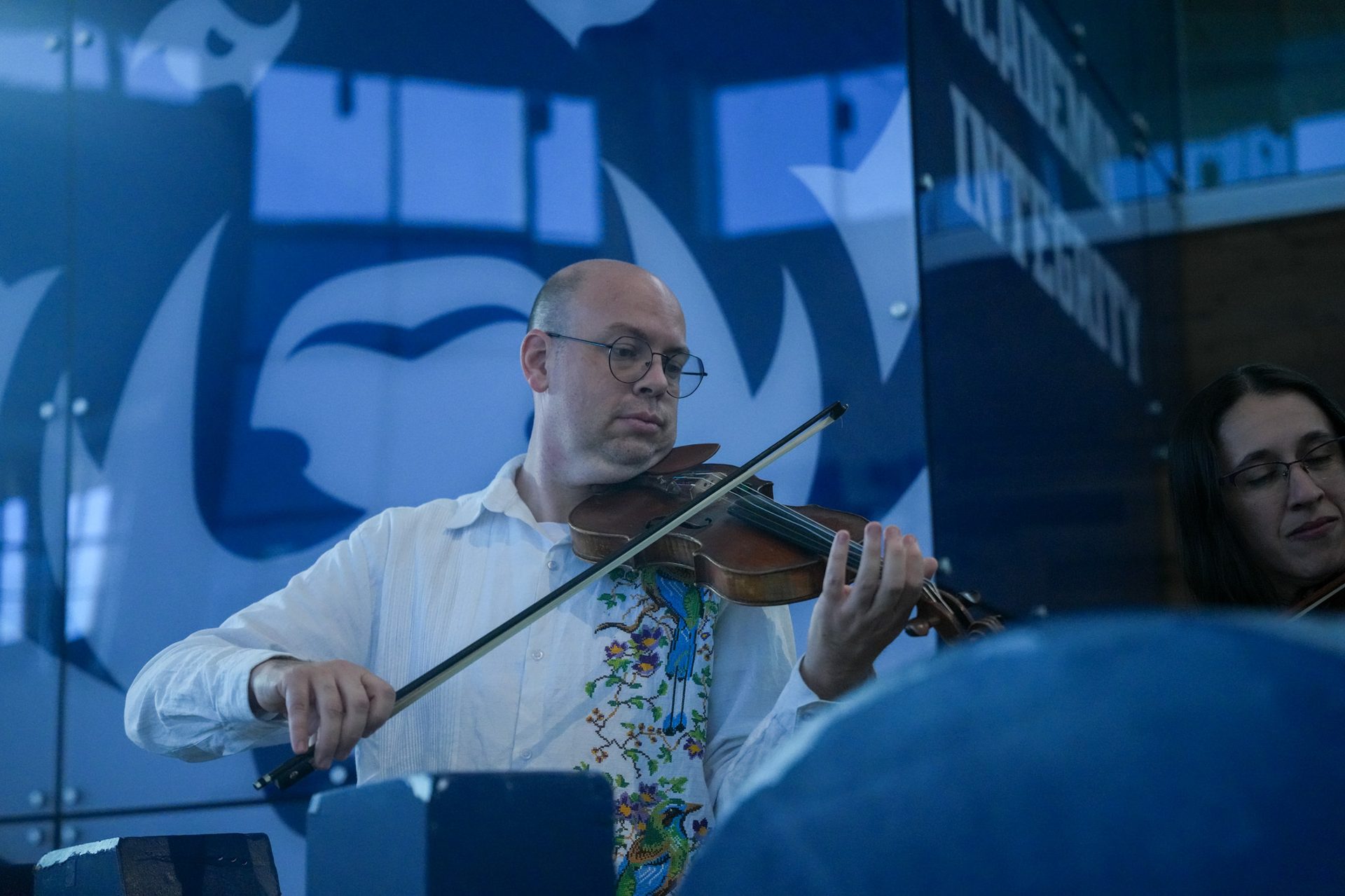 Bald man in glasses playing a violin, wearing an embroidered shirt. Another musician is partially visible.