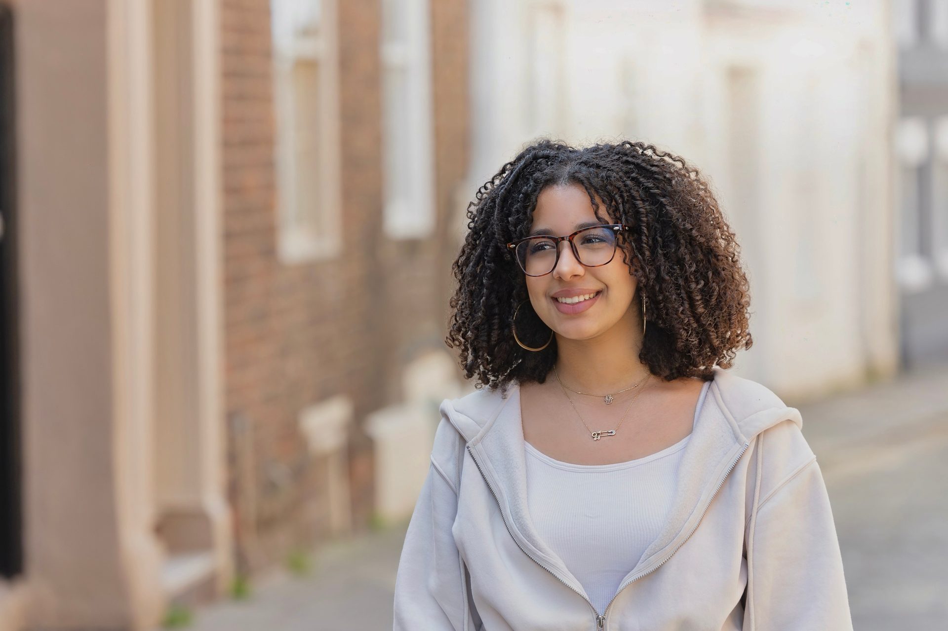 A smiling young woman with curly hair and glasses standing outdoors.