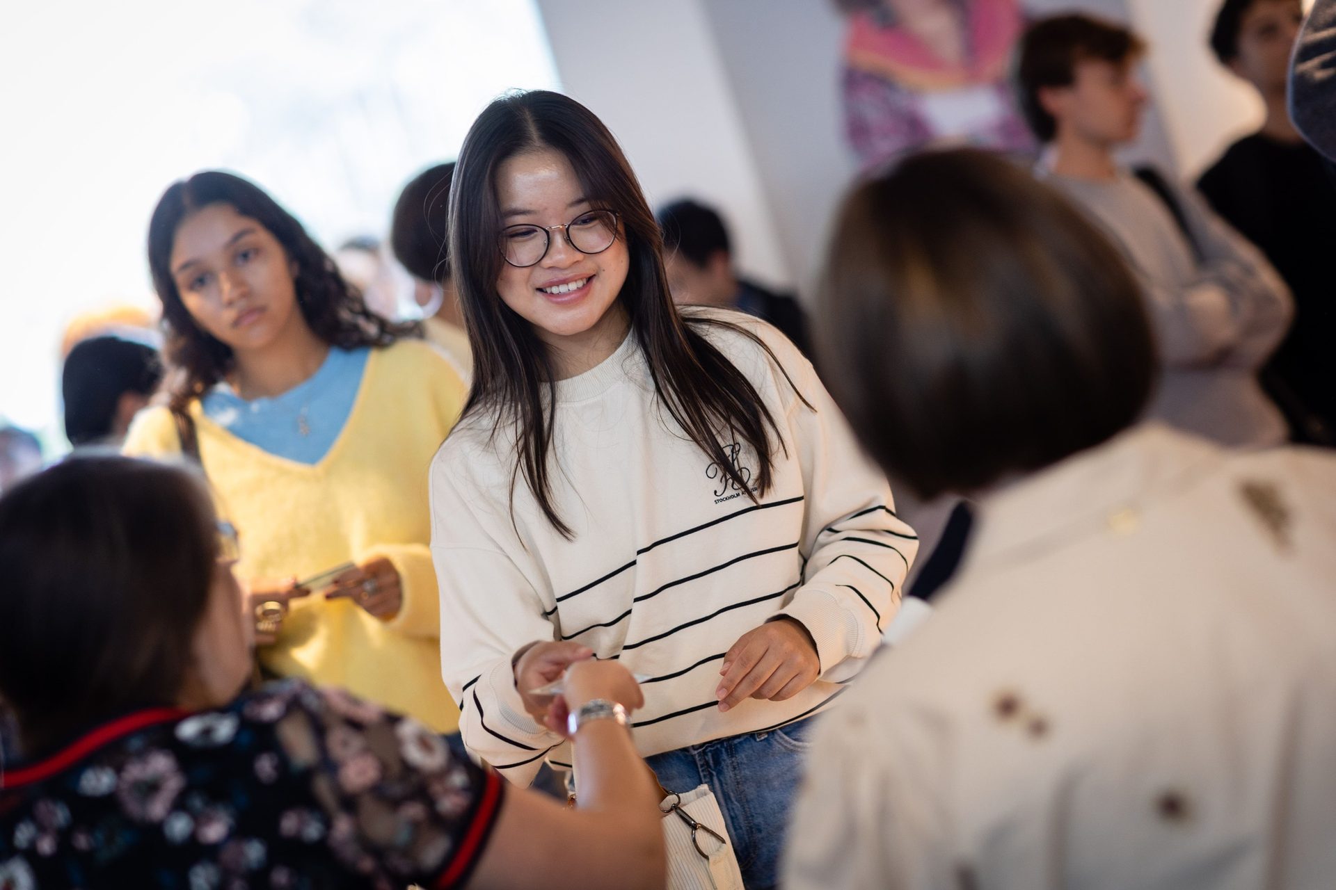 Smiling young Asian woman in glasses and striped sweater exchanging items with another person.