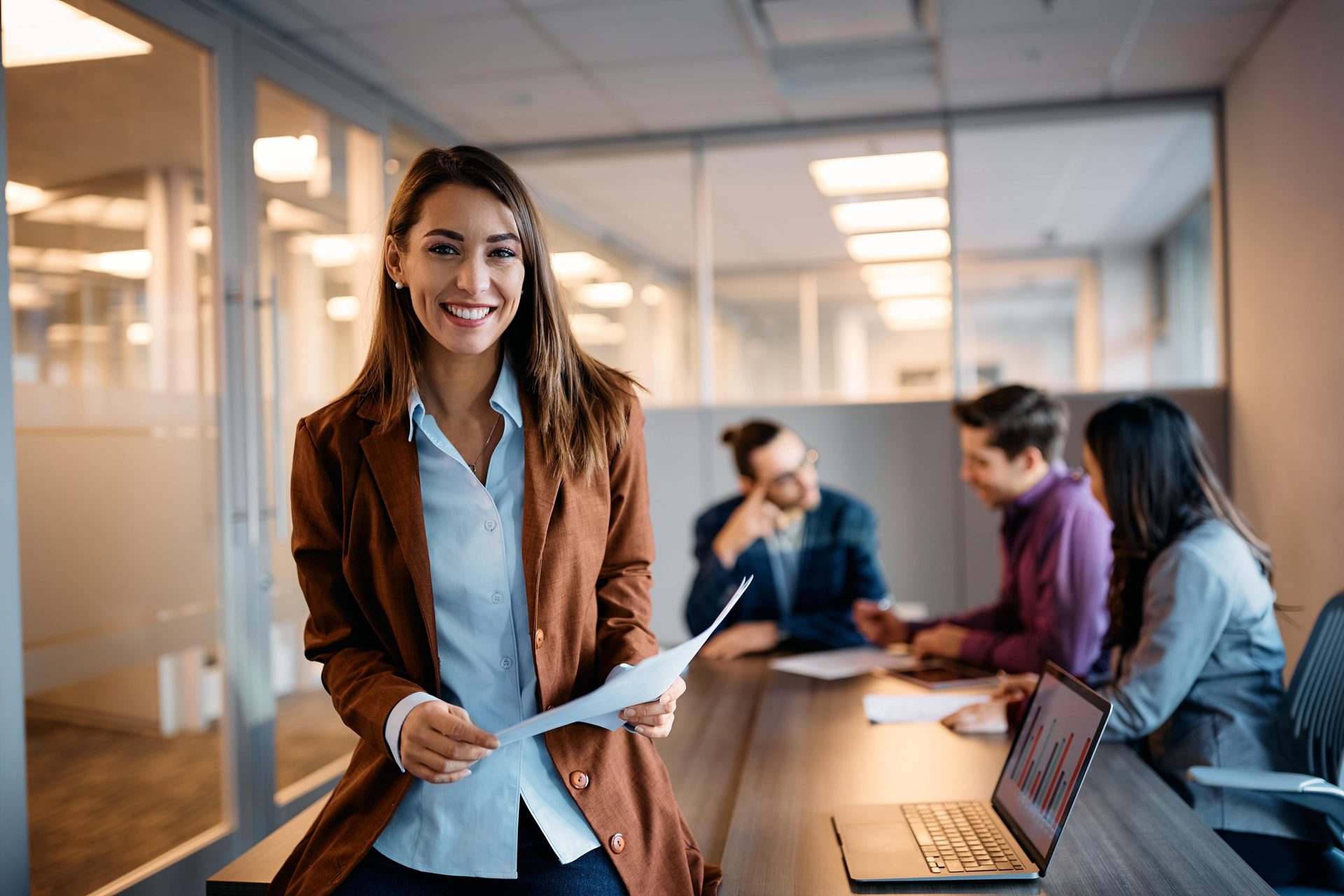 Smiling woman in an office holding papers, with blurred colleagues in the background.
