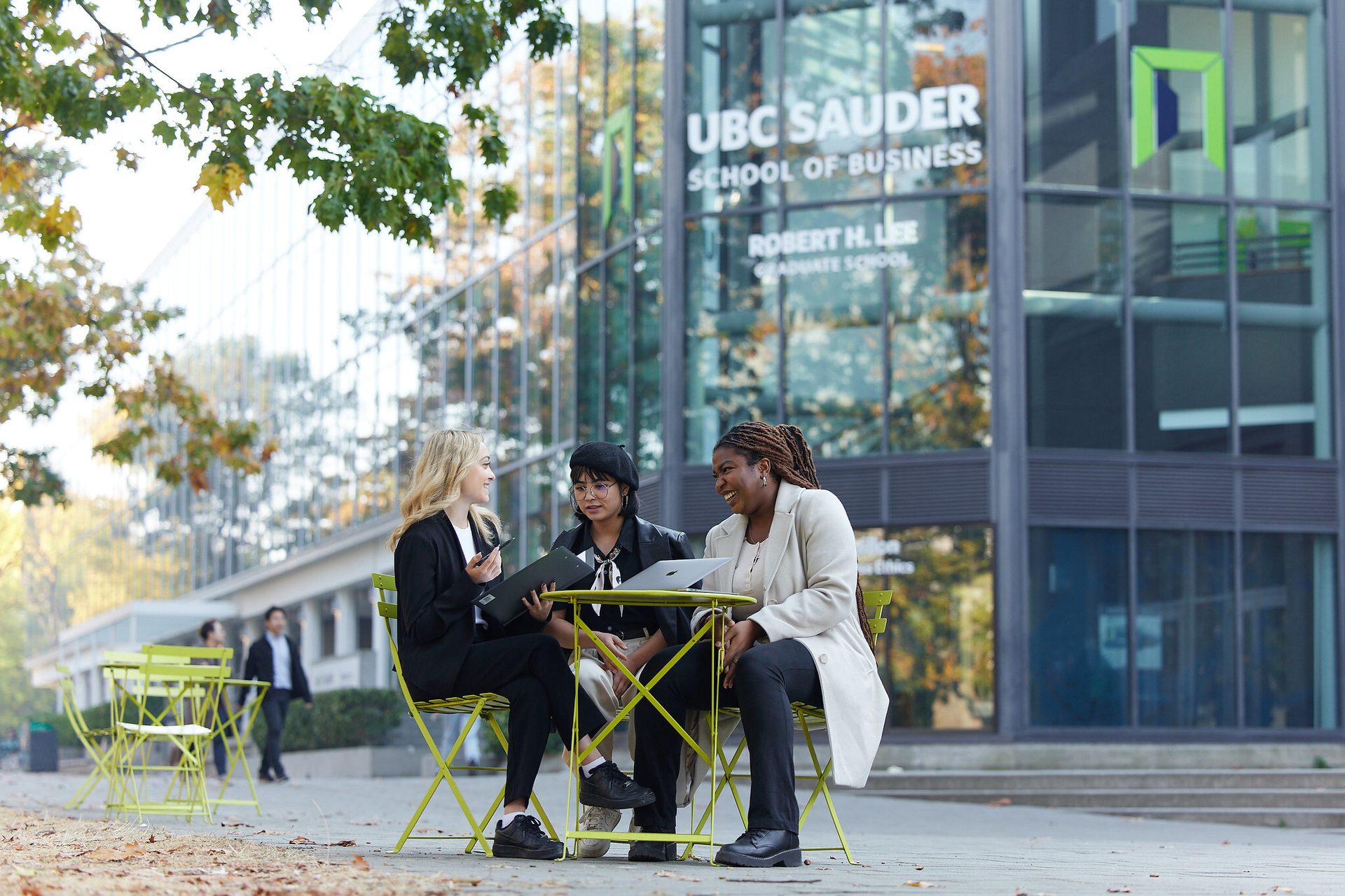 Three diverse women at an outdoor table with the UBC Sauder School of Business building behind them.