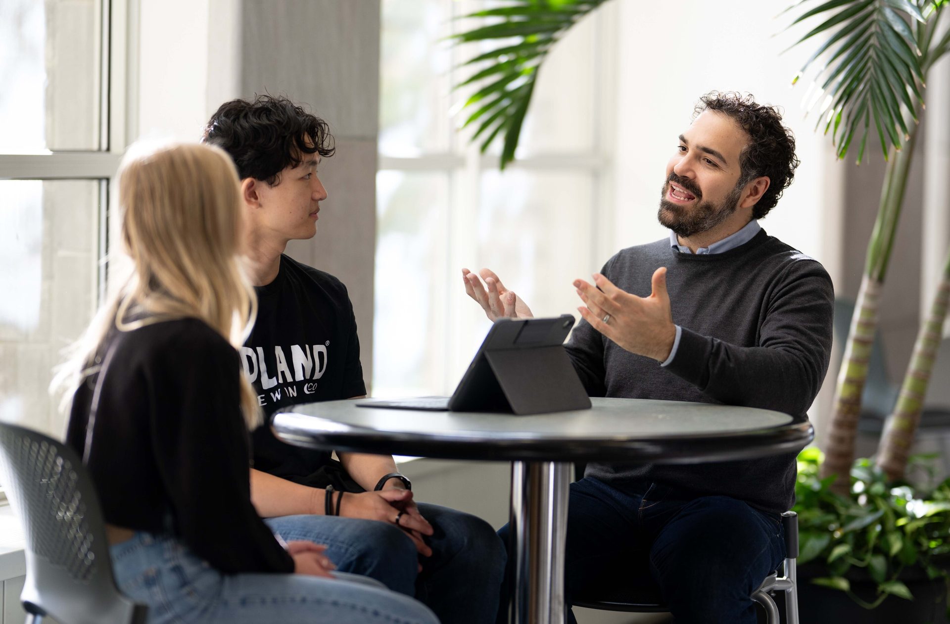 A bearded man speaks, gesturing, to two listeners at a table with a tablet.