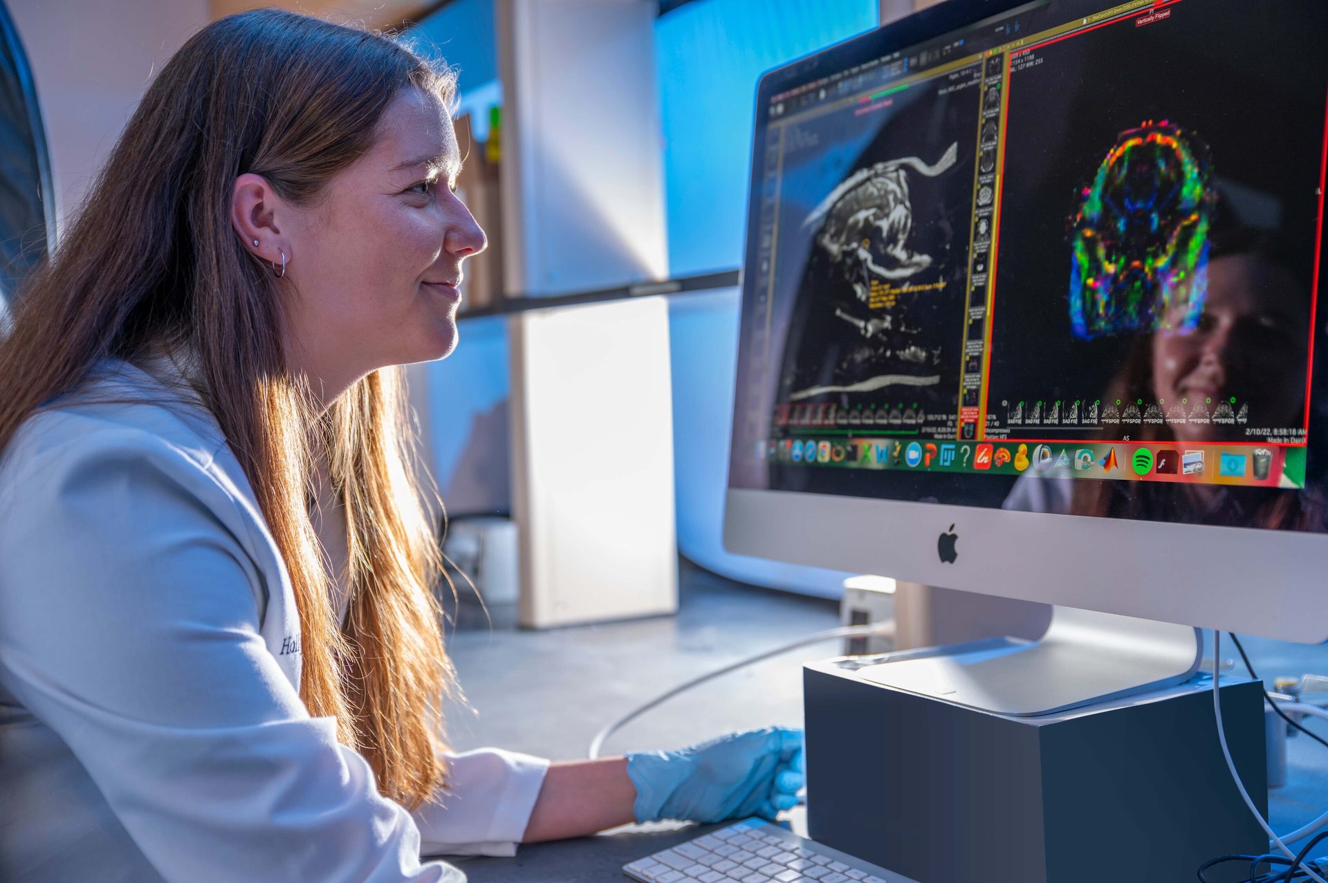 Woman in lab coat views medical scans on a computer monitor, her reflection visible.