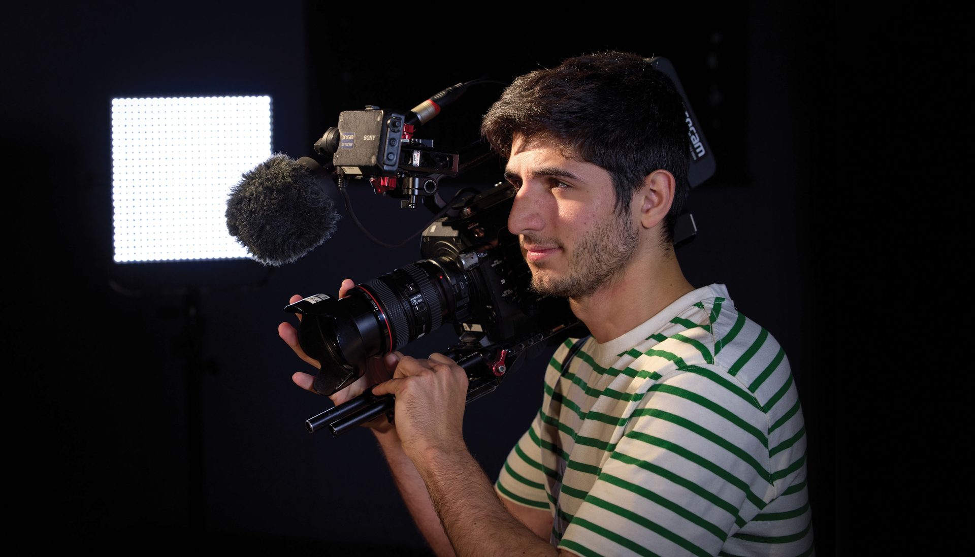 Young man operating a professional video camera rig, lit by a bright LED panel.