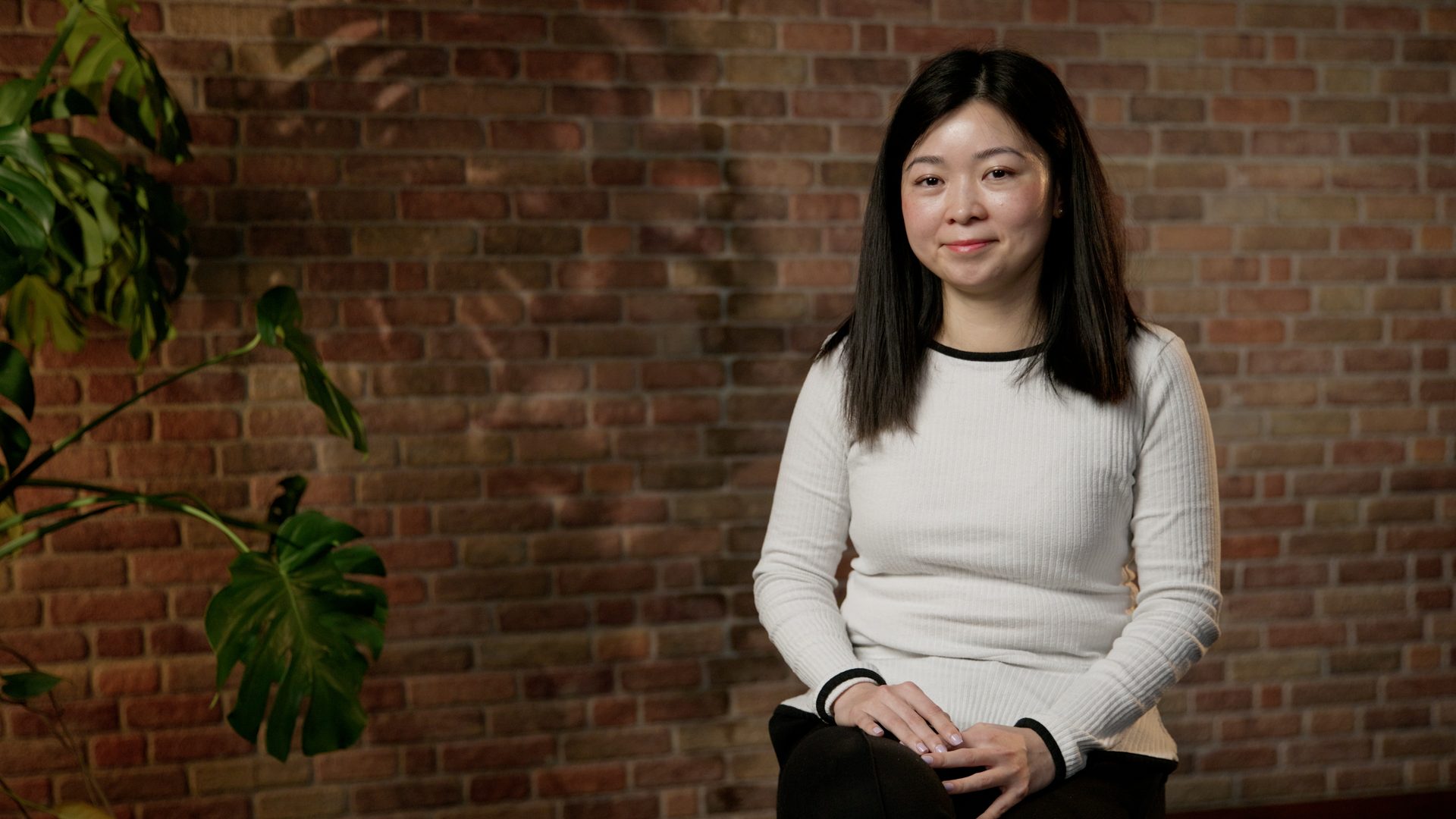 Smiling woman in white top, sitting against a brick wall with a plant.