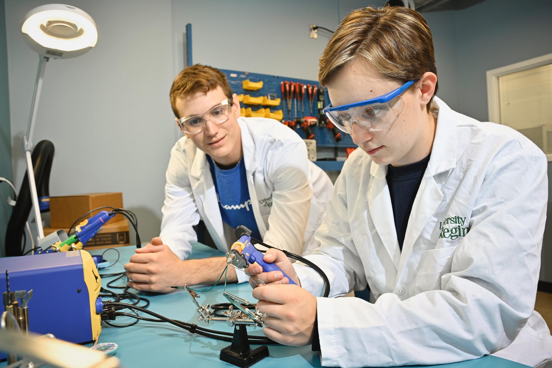 Two students in lab coats & safety glasses: one solders electronics, the other watches.