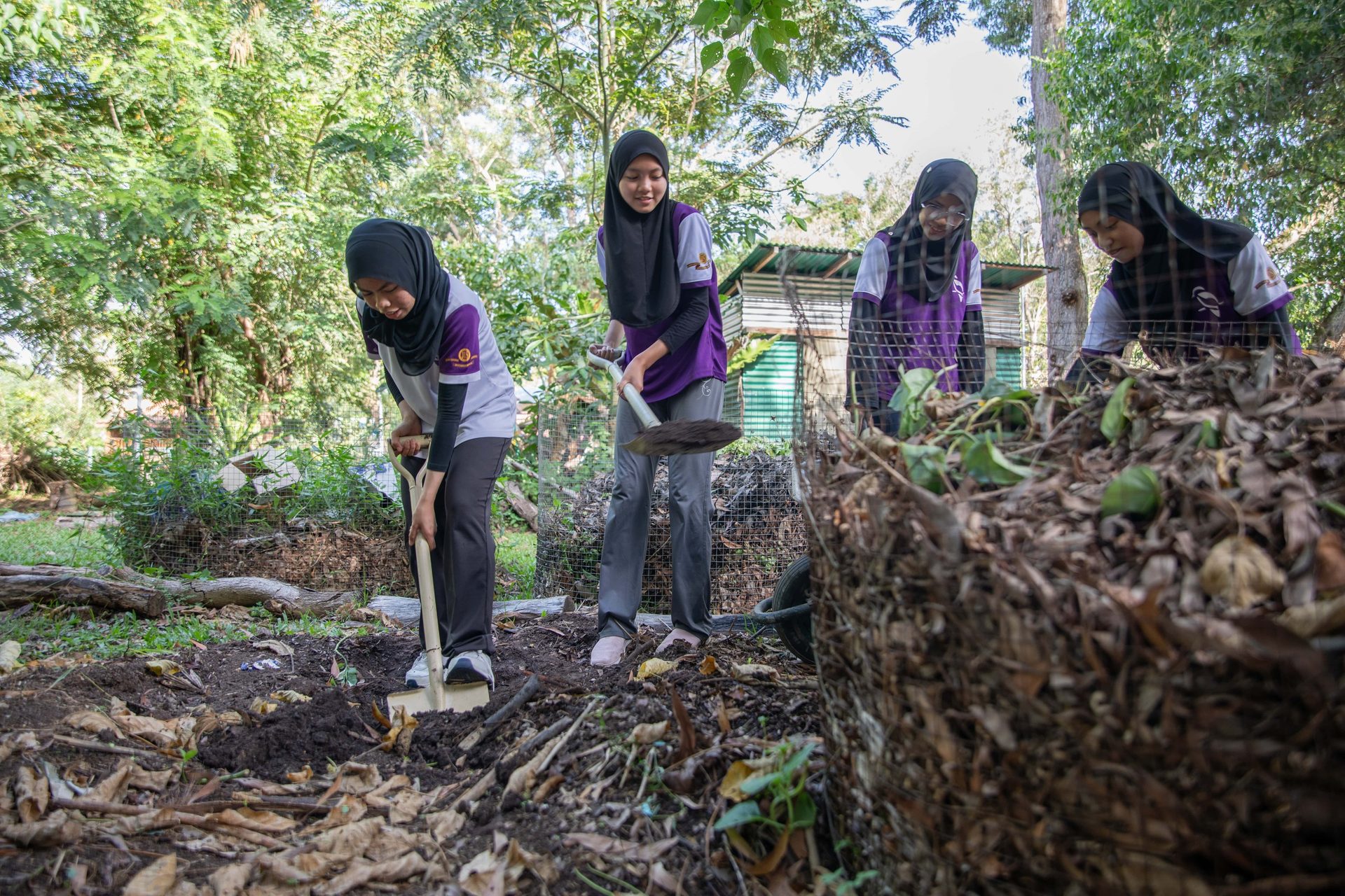 Four women in hijabs tending to compost and digging in a garden.