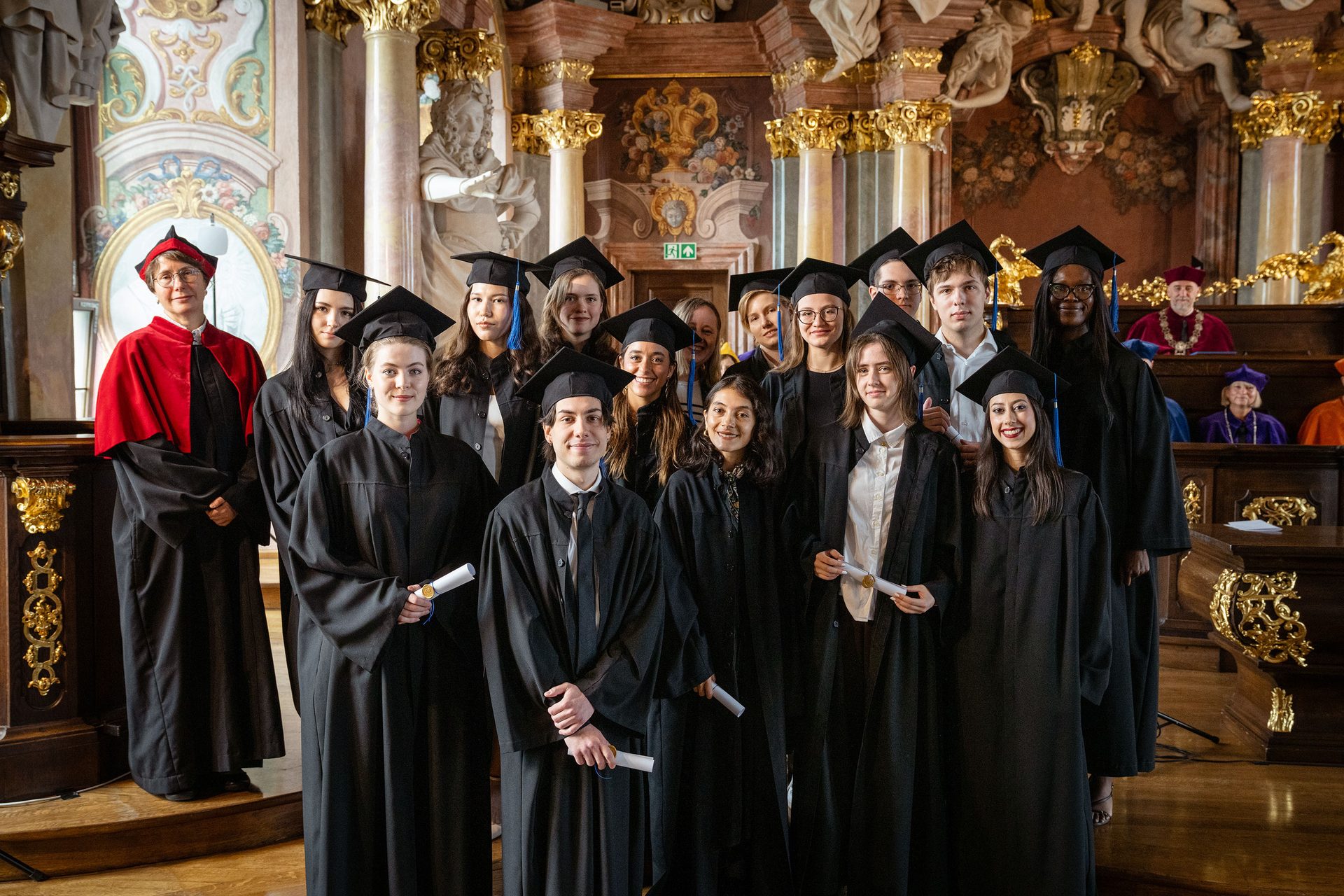 Diverse graduates in academic regalia with faculty, holding diplomas in a grand hall.