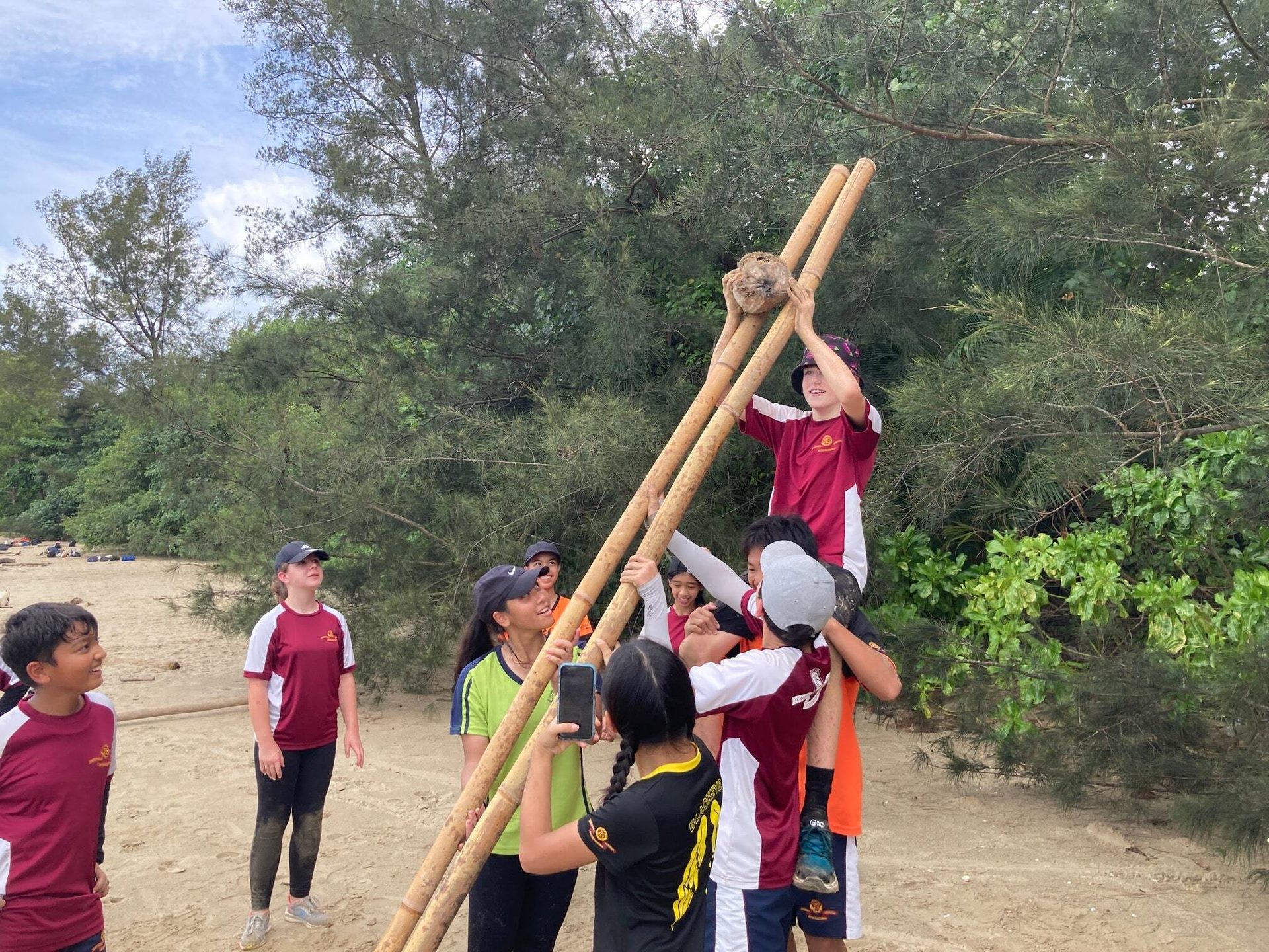 Youth on beach team-lift coconut with bamboo poles; one photographs.