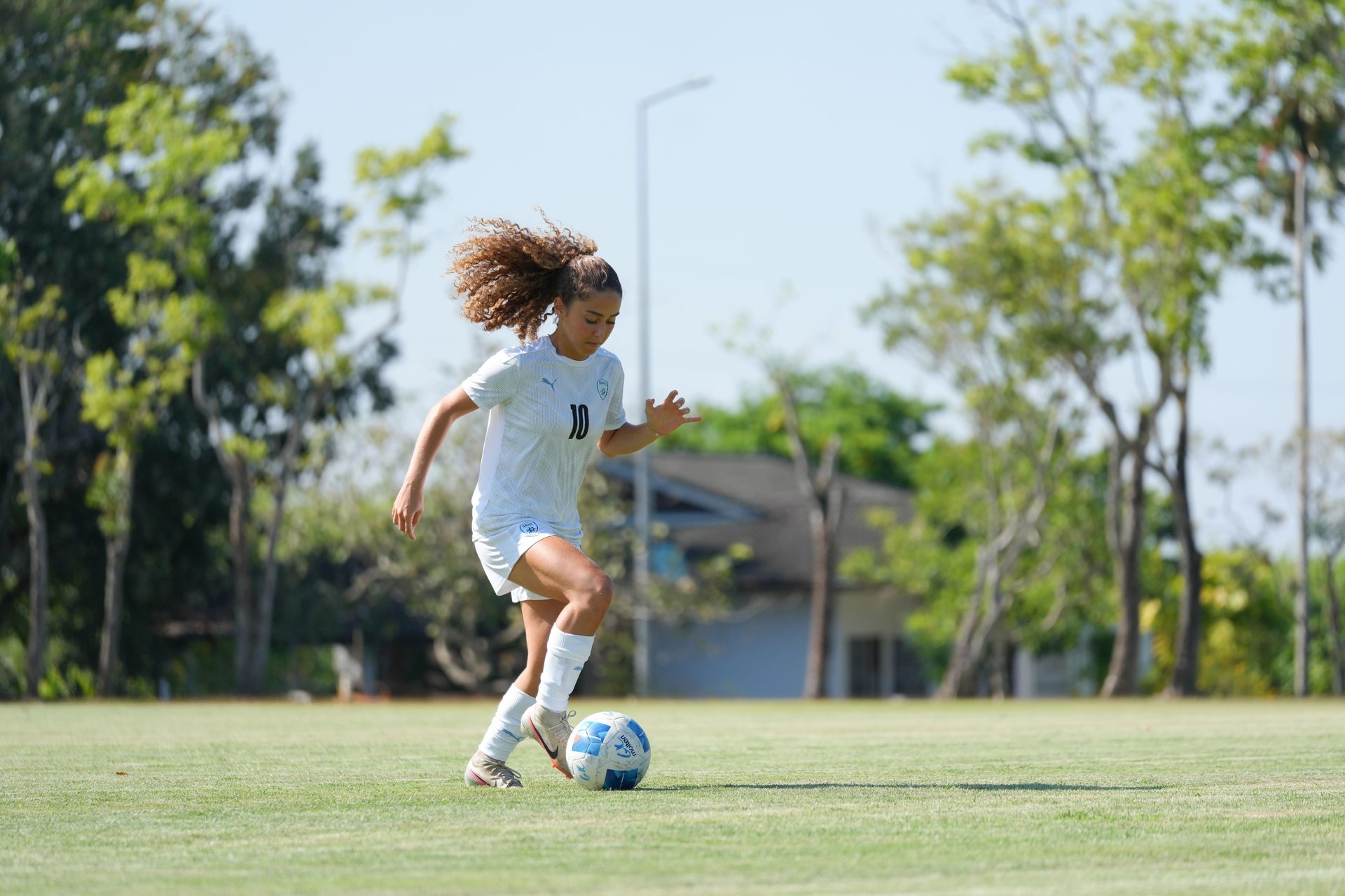 Young female soccer player with curly hair dribbling a ball on a sunny green field.
