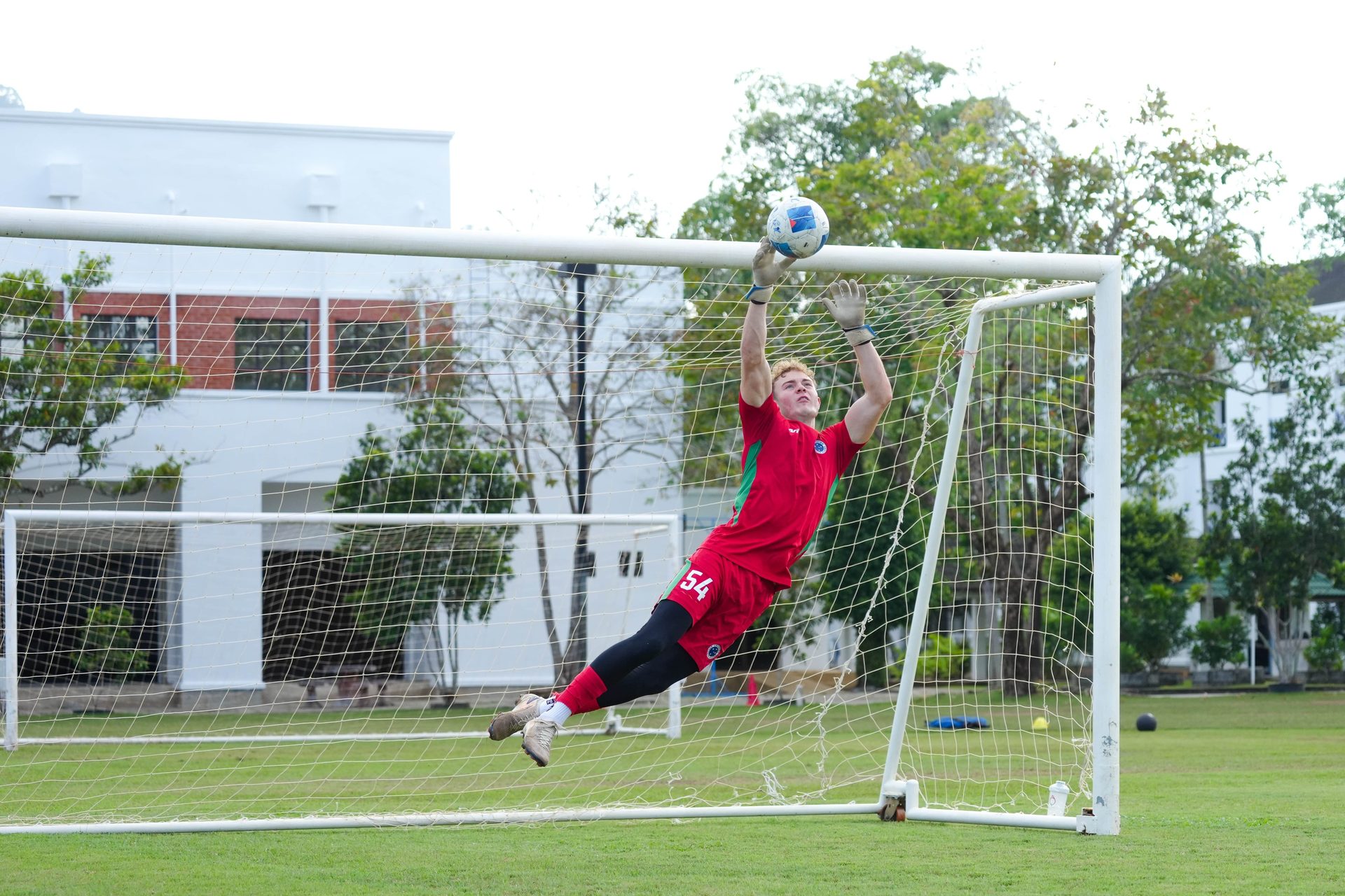 Male soccer goalkeeper in red jersey diving to catch a ball near the goal's crossbar.