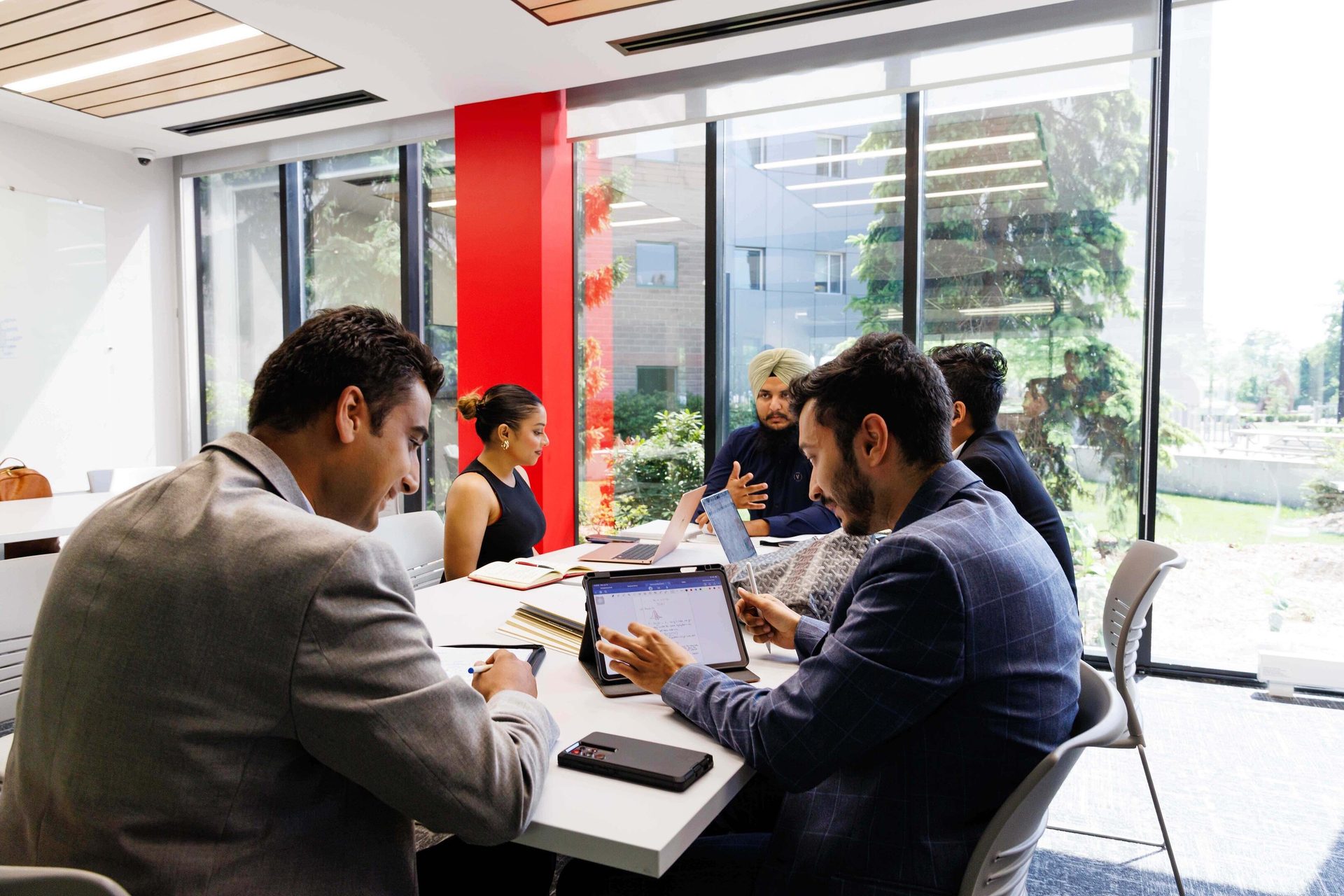Five people in a modern meeting room, collaborating with laptops and tablets.